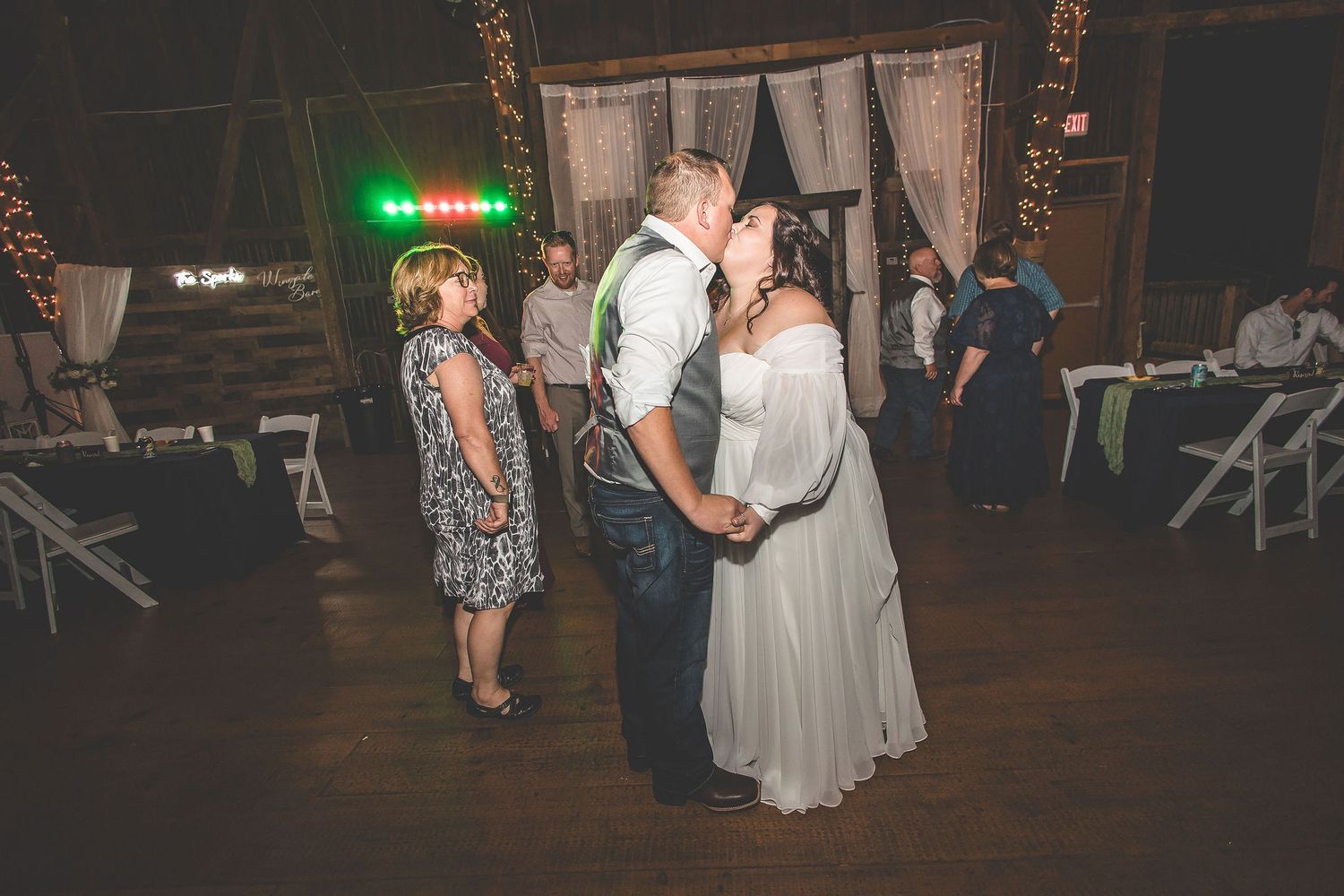 Evening wedding reception with guests dancing under string lights in a rustic barn setting.