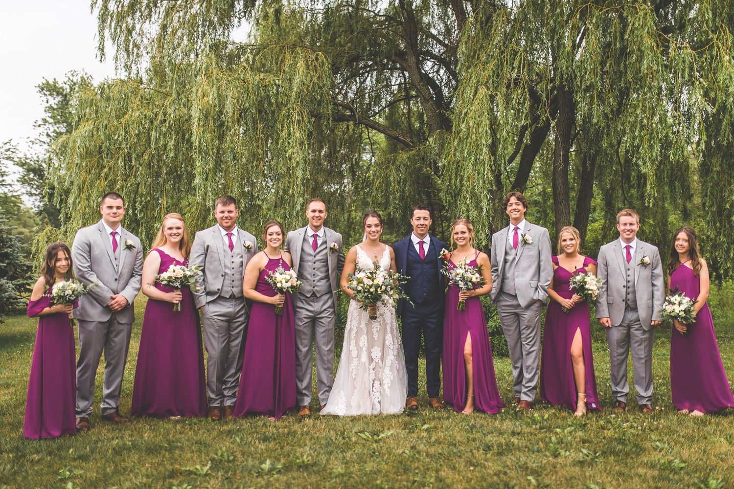 Wedding party poses under a weeping willow tree wearing purple and gray formal attire.