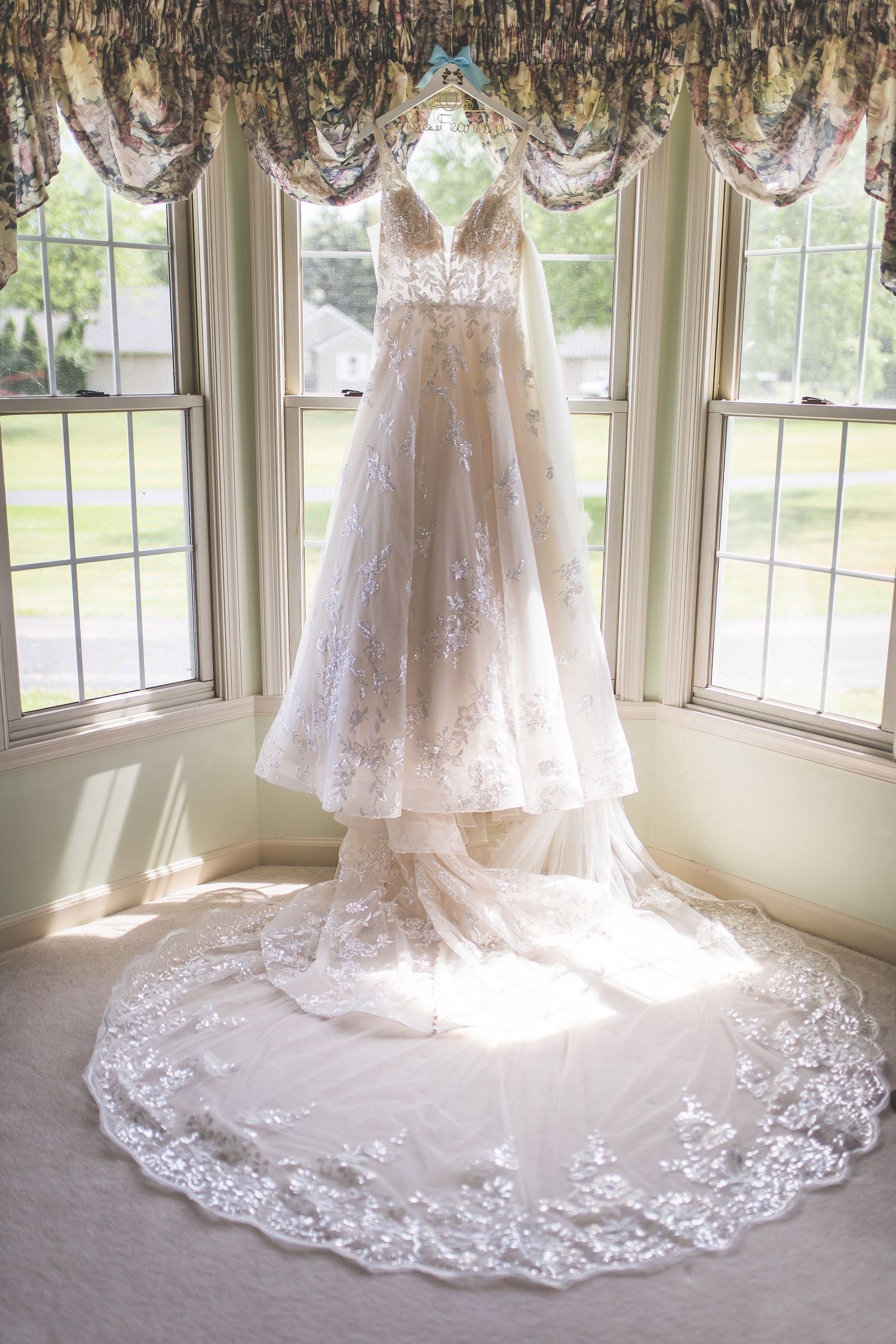 Wedding dress with intricate lace train displayed in sunlit window setting.