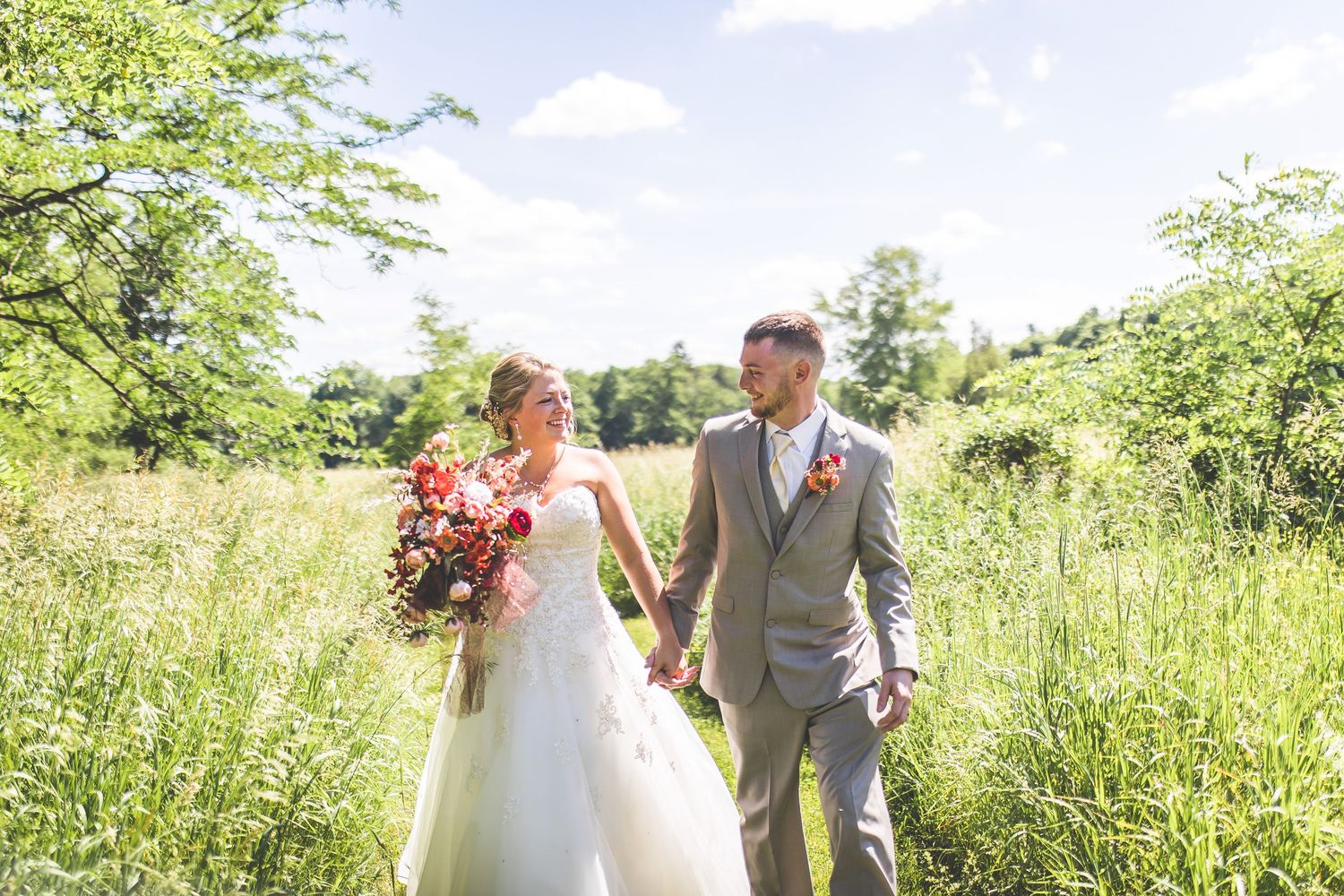 A couple walks hand in hand through a sunlit meadow on their wedding day.