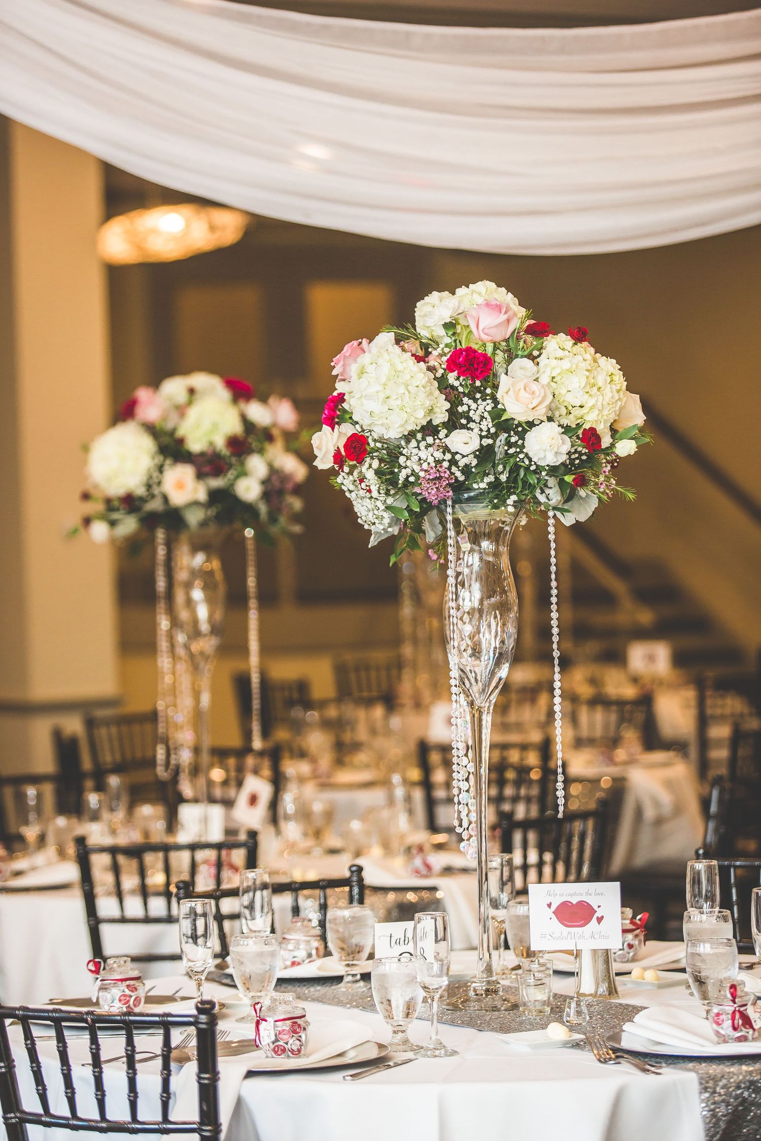 Elegant wedding reception centerpieces with tall crystal vases filled with white hydrangeas and roses.