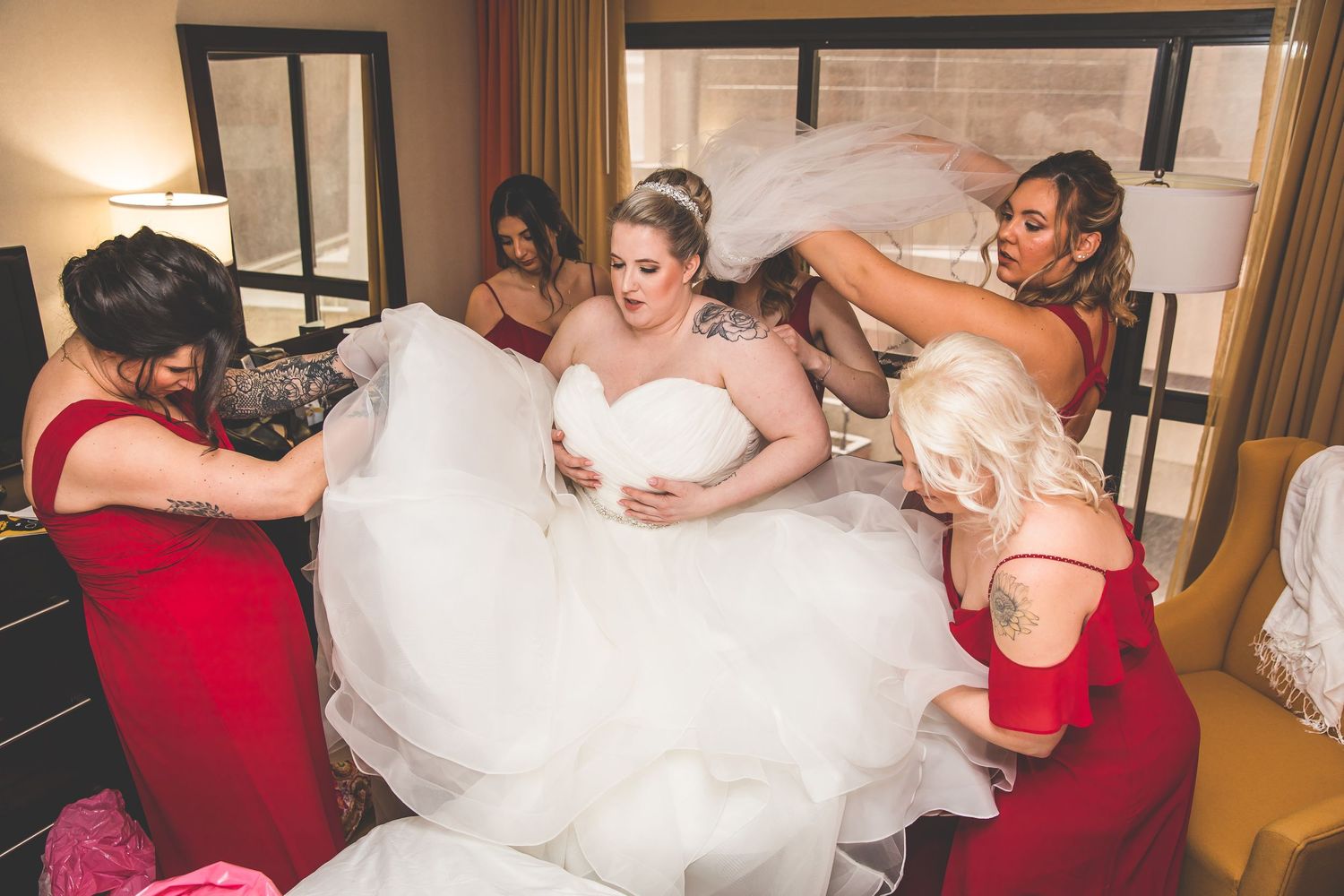Wedding party wearing red dresses and white gown embracing in a hotel room.