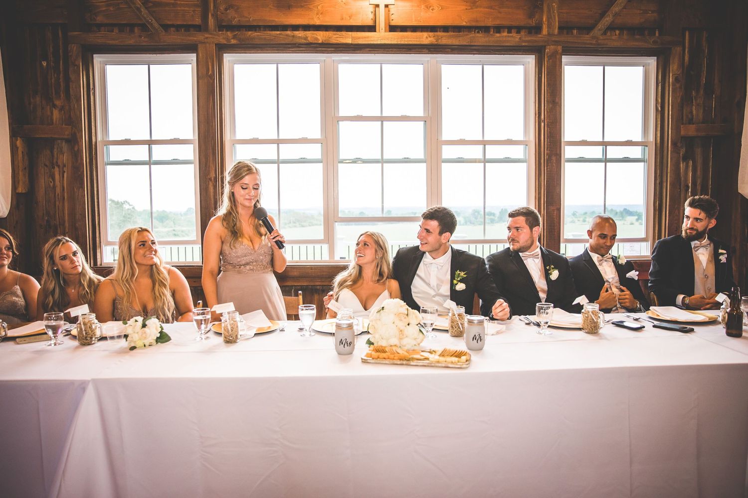 Wedding reception speech being delivered at long white-clothed table with guests seated.