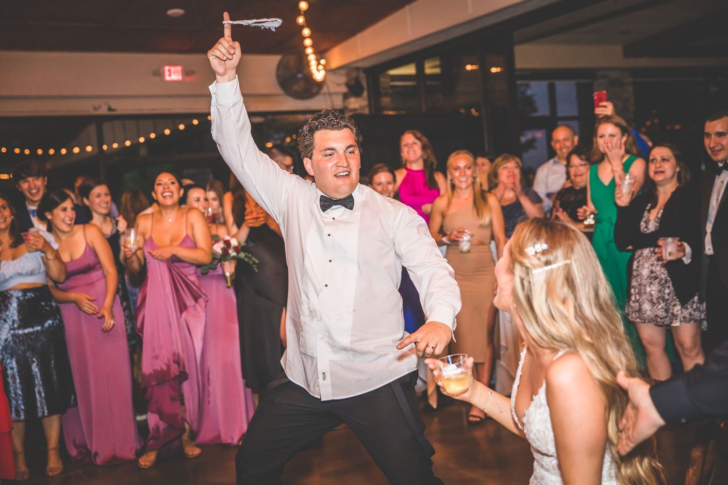 Wedding party dancing and celebrating at reception under decorative string lights.