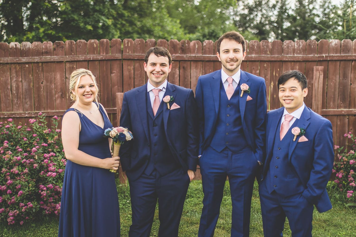 Groomsmen in matching navy suits with orange boutonnieres pose against wooden fence.