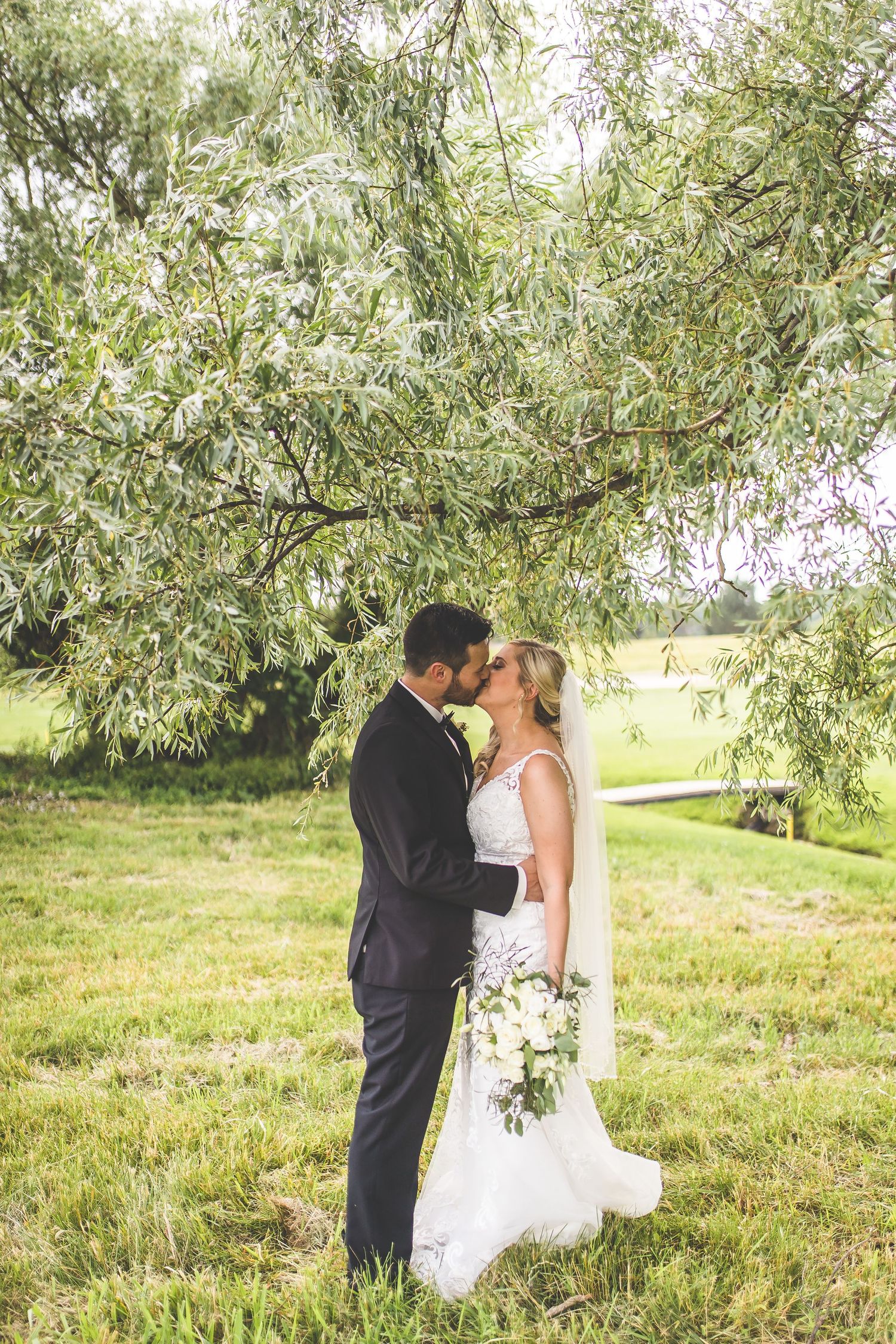 Bride and groom share an intimate moment under a willow tree in a serene outdoor setting.