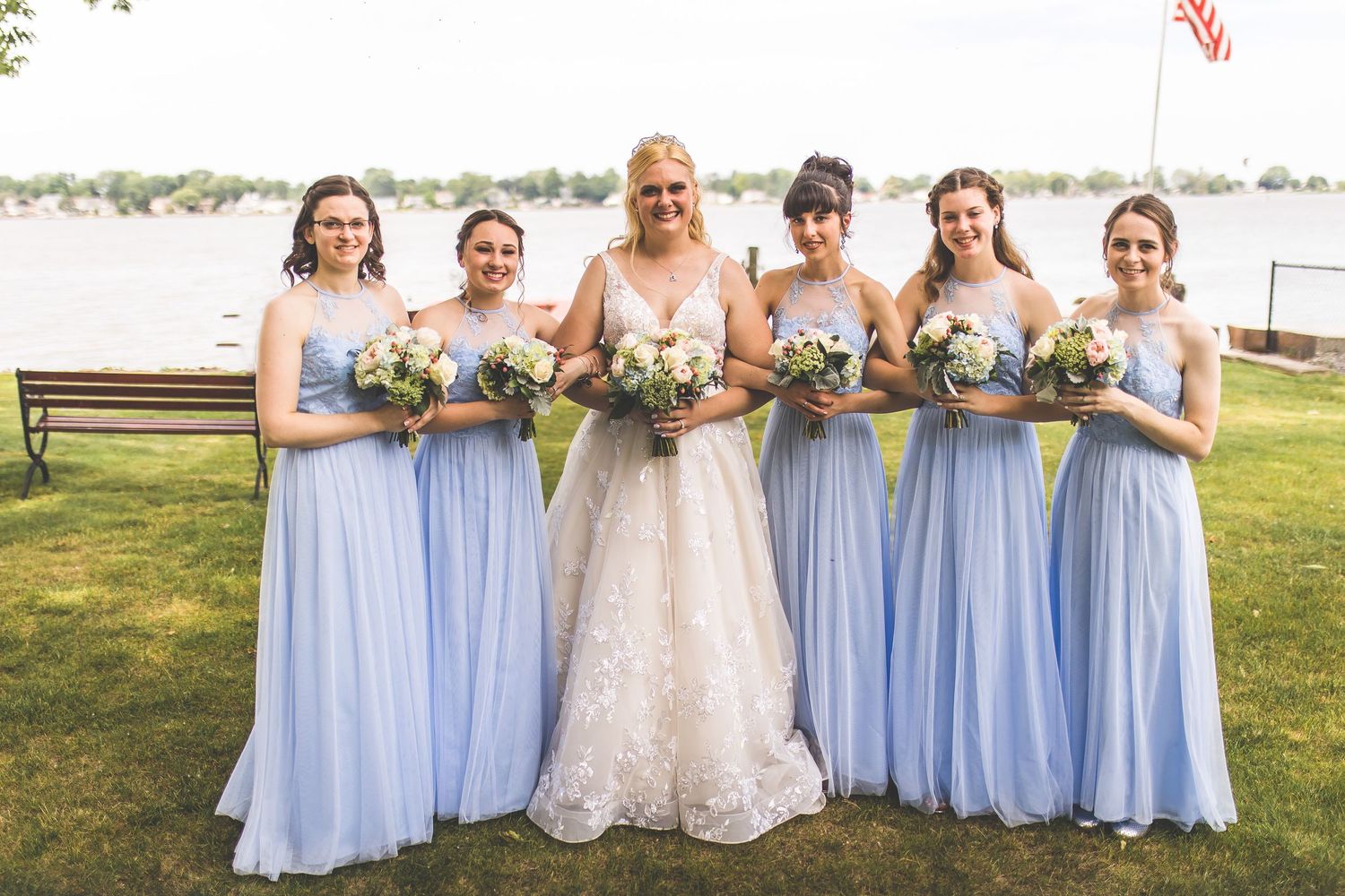 Bridesmaids in light blue dresses stand together with bride in white gown outdoors.