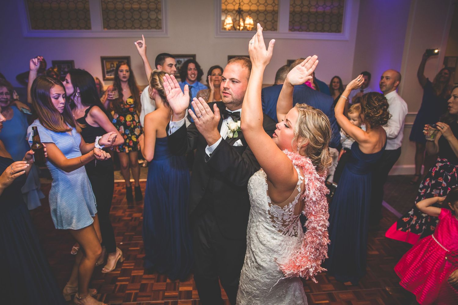 Wedding party dancing energetically under purple mood lighting at reception.