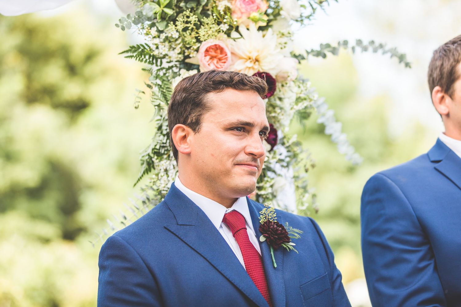 Groomsmen in navy suits with red ties stand together outdoors.