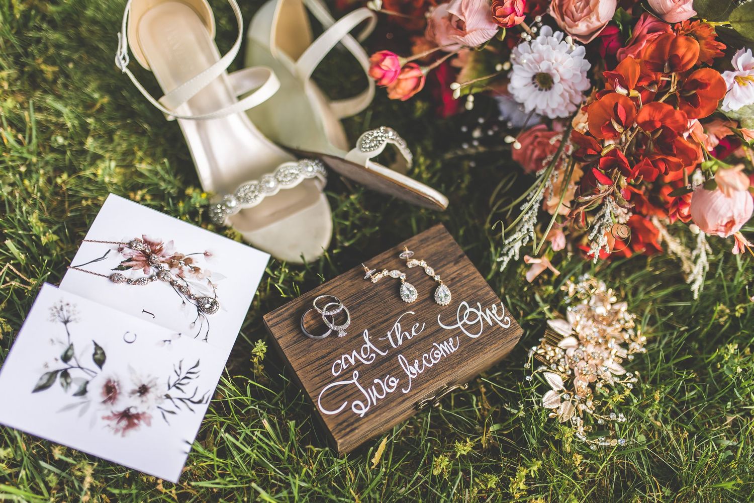 Wedding detail flatlay with floral arrangements, shoes, and wooden signs on grass.