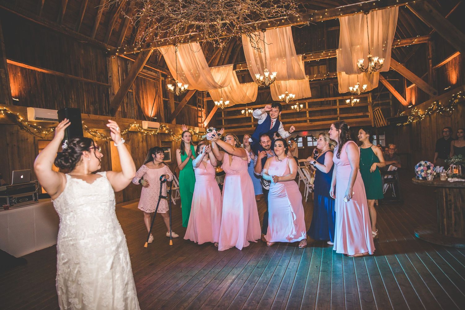 Wedding reception dance floor filled with guests in colorful dresses under warm lighting in a rustic barn venue.
