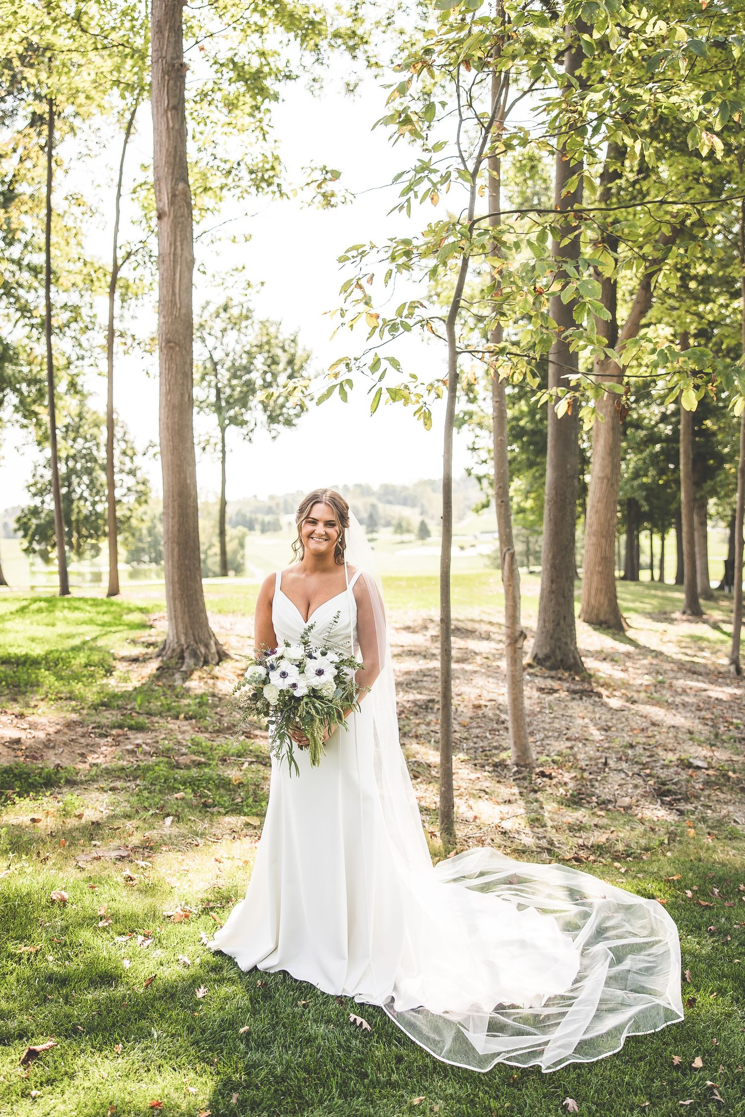 White wedding dress with flowing train captured in sunlit wooded outdoor setting.