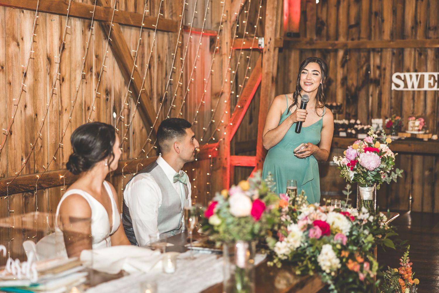 A wedding toast being given during dinner in a rustic barn venue with floral decorations.