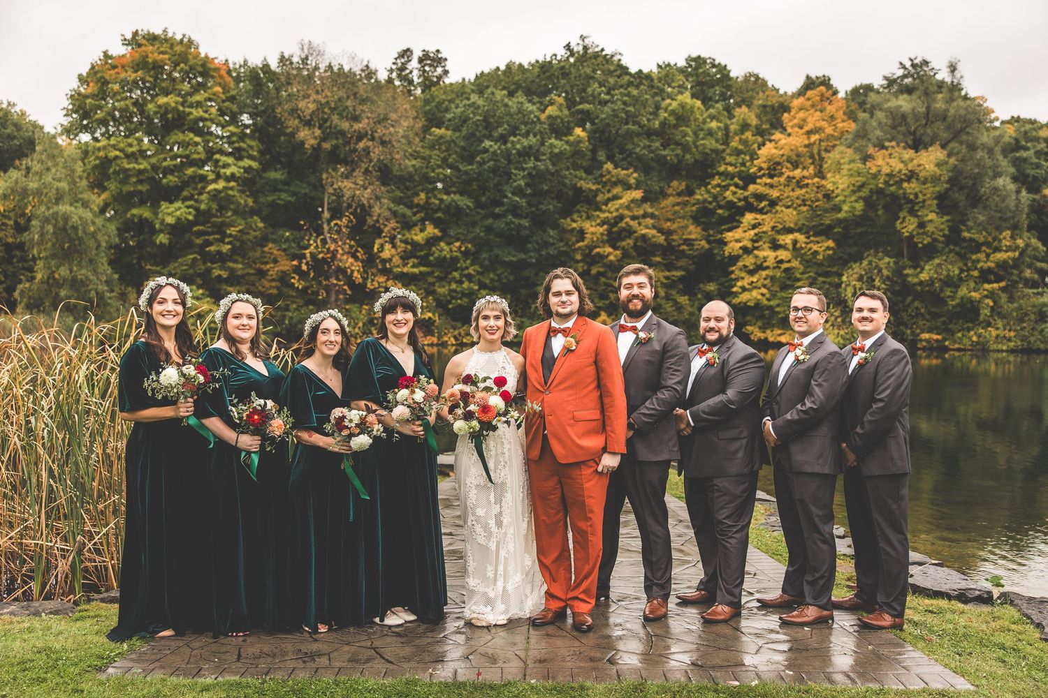 Wedding party in black and formal attire poses by lake with autumn trees in background.