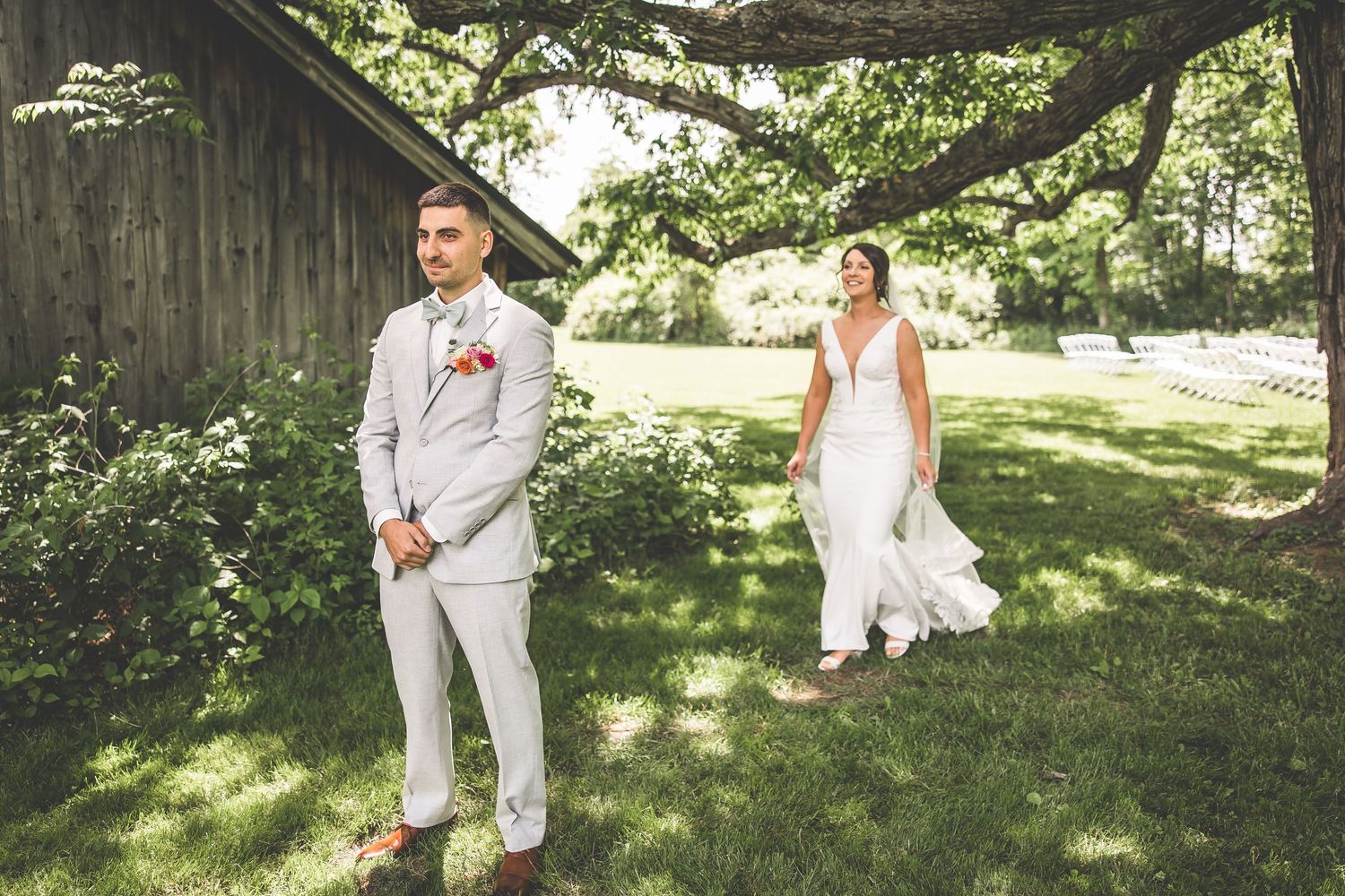 Wedding couple poses in front of rustic barn with green foliage.