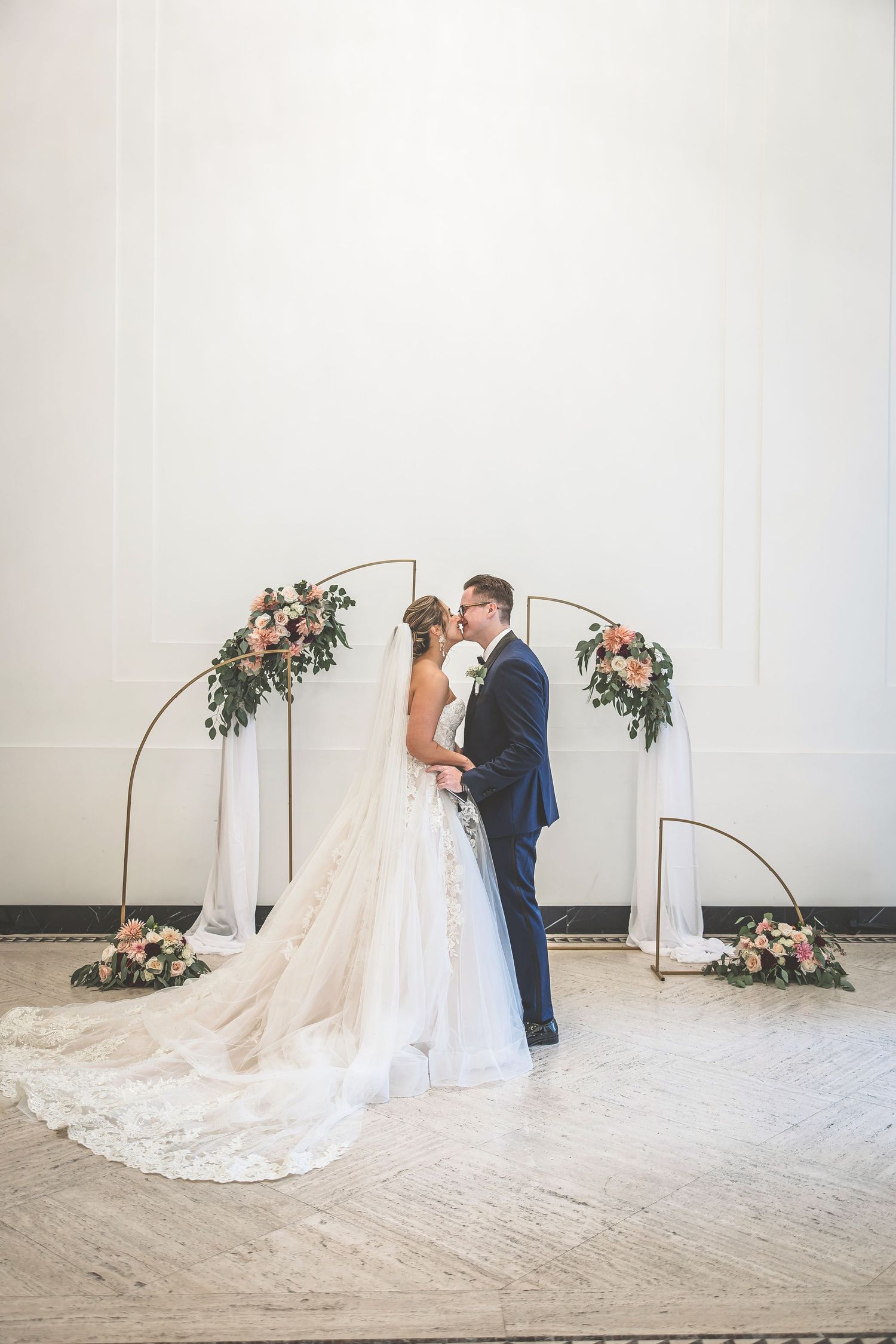 A wedding couple poses under a modern arch decorated with pink roses and greenery.