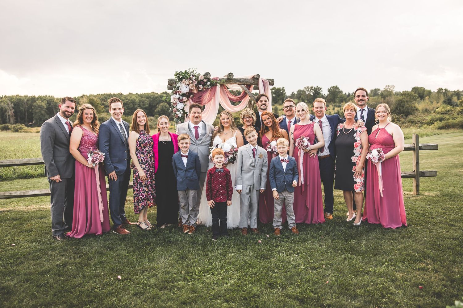 Large wedding party group photo with pink and gray color scheme taken in a grassy outdoor setting.