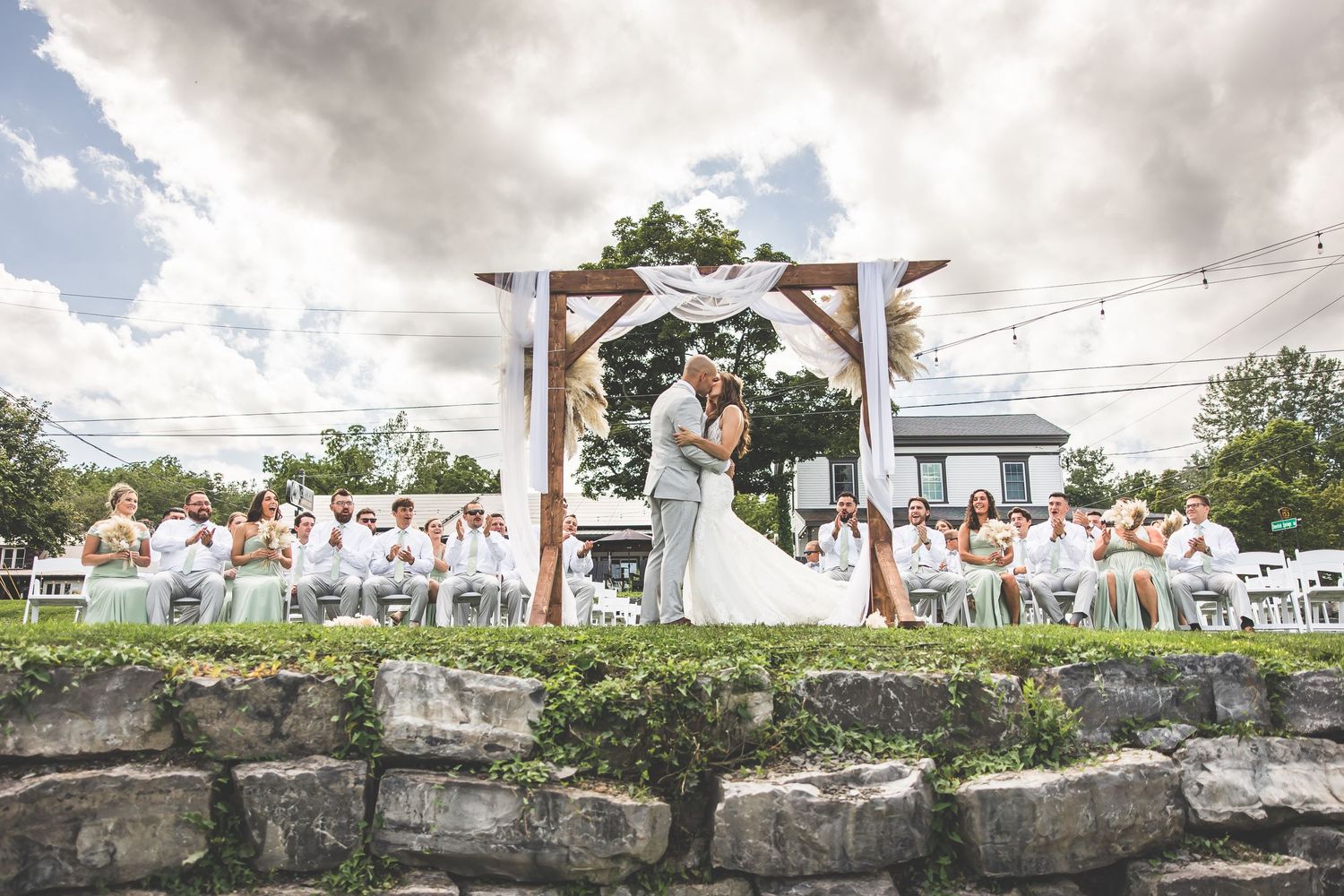 Wedding ceremony under wooden arch with string lights and stone wall landscaping.
