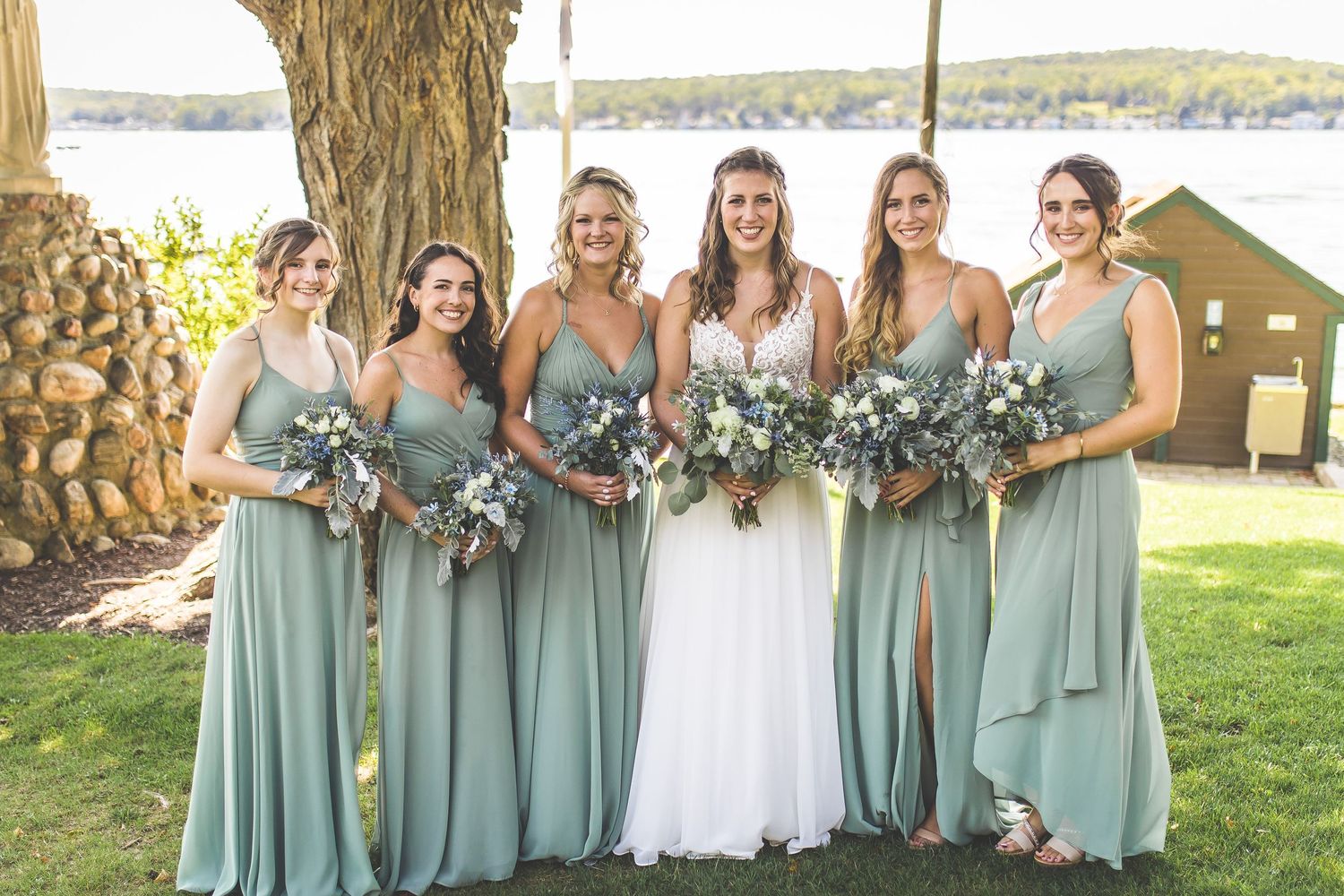 Bridesmaids in matching sage green dresses pose with bouquets by lakeside venue.
