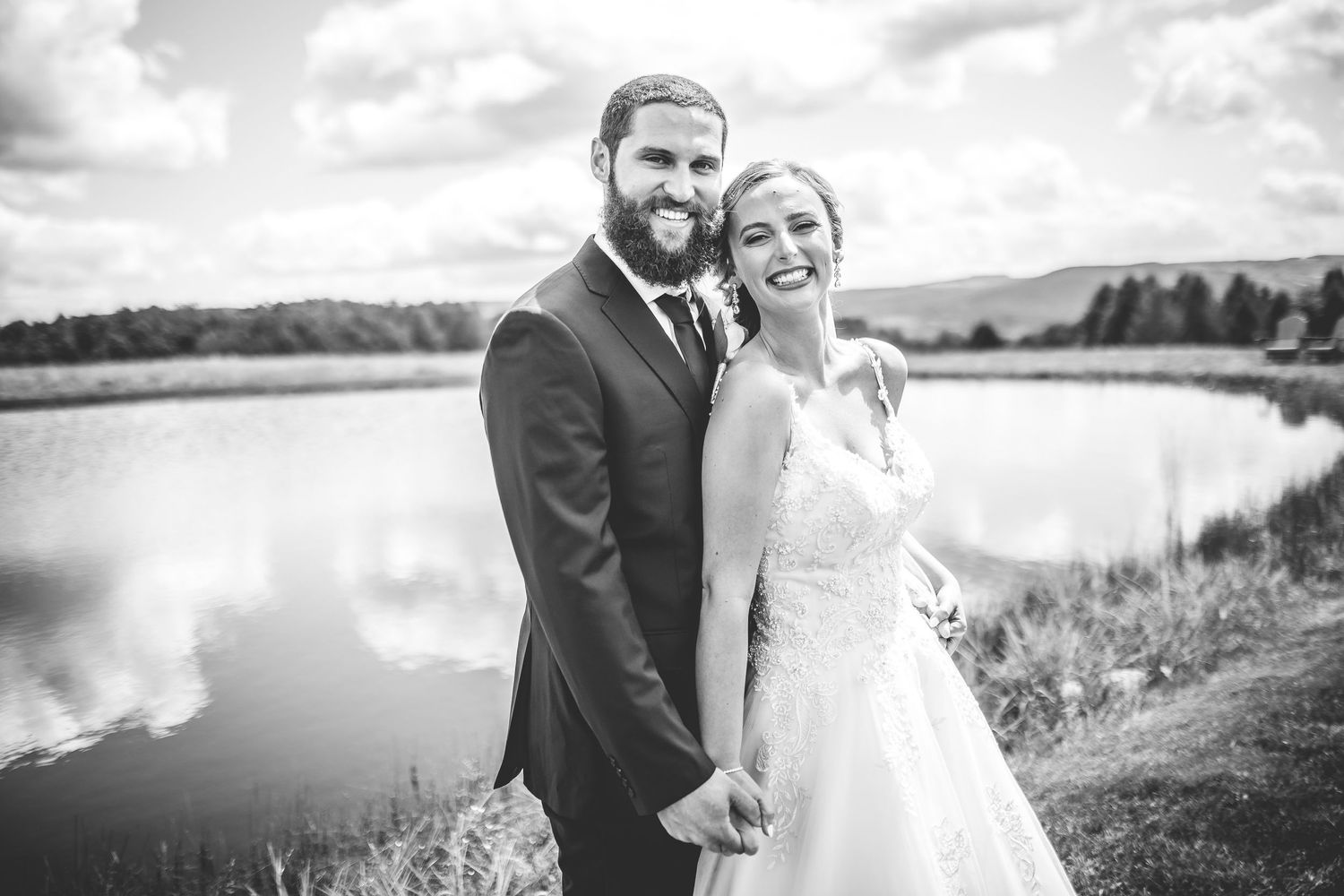 Black and white portrait of newlyweds by a lake with rolling hills in background.