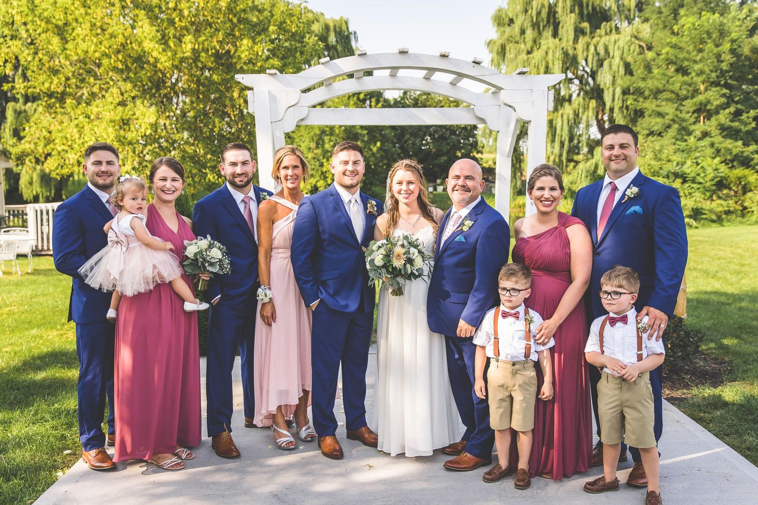 Extended wedding party and family members gather for formal photos under a white garden arbor.