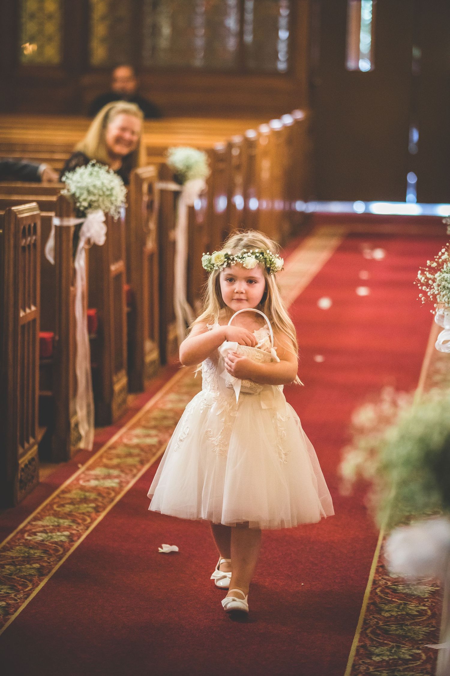 Flower girls in white tulle dresses walk down red-carpeted church aisle.