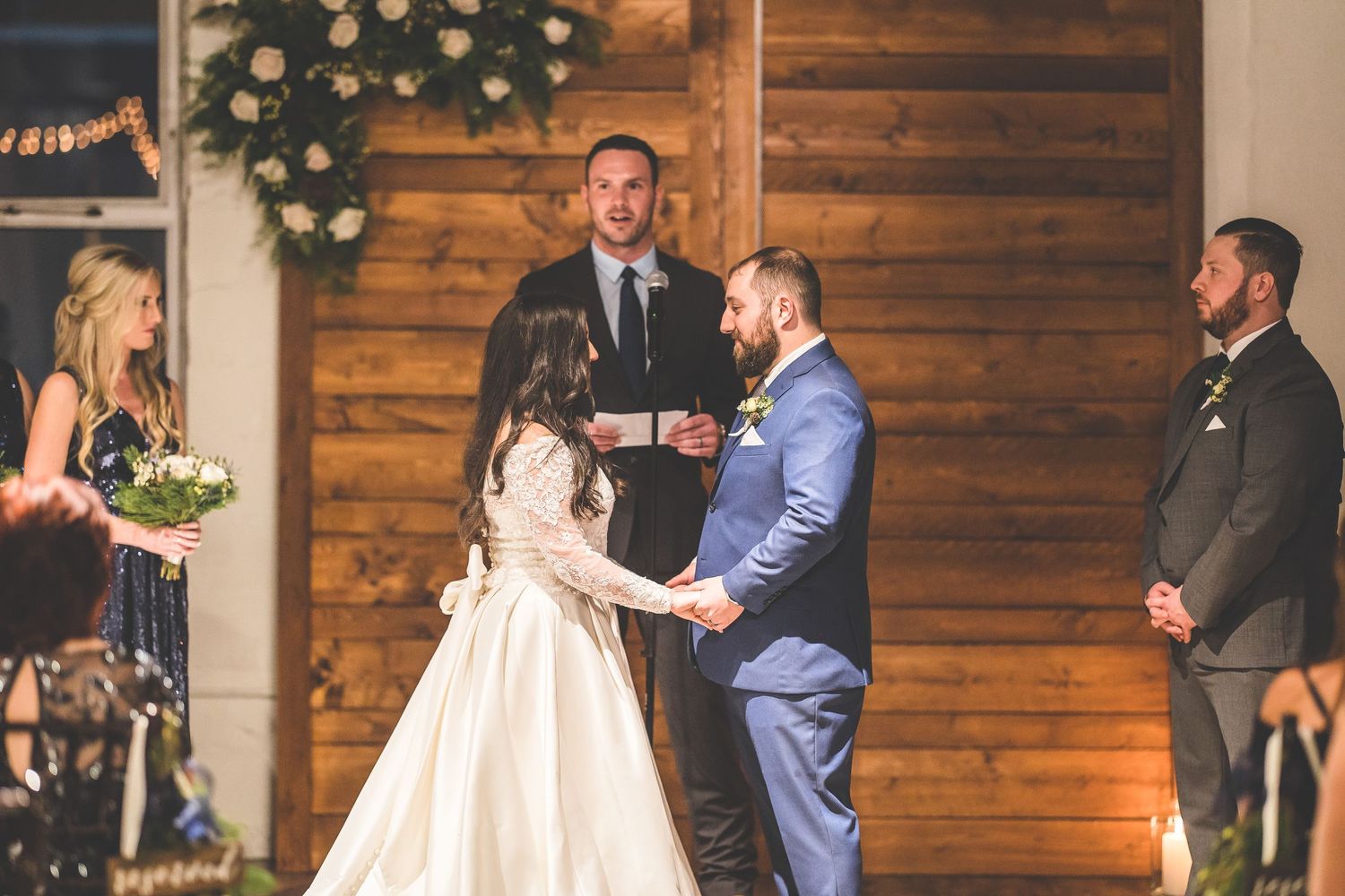 Wedding party gathered in front of a rustic wooden wall decorated with floral arrangements.