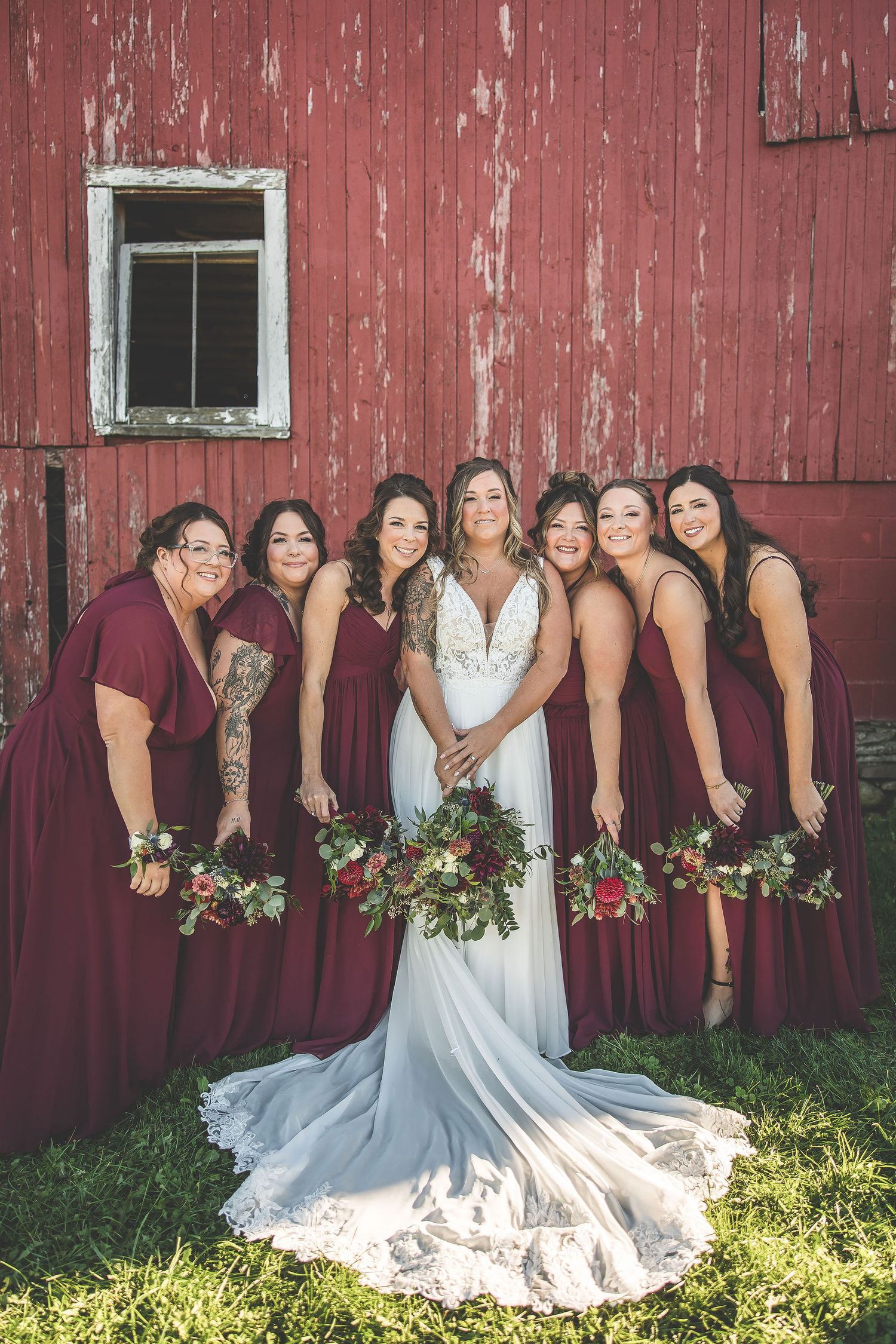 Wedding party members line up against weathered red barn wall in burgundy and white attire.