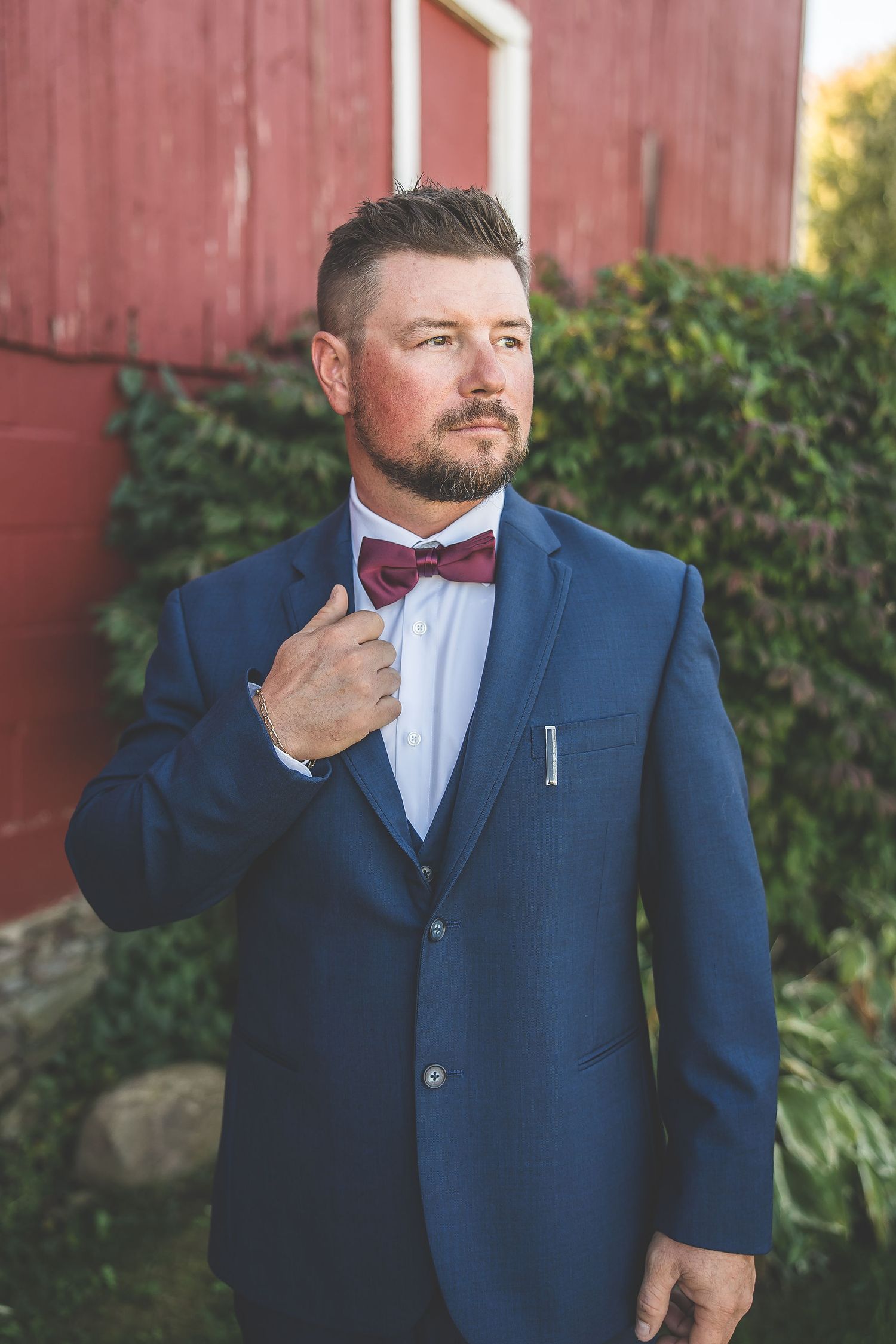 Groom adjusts burgundy bowtie while posing against rustic barn backdrop.