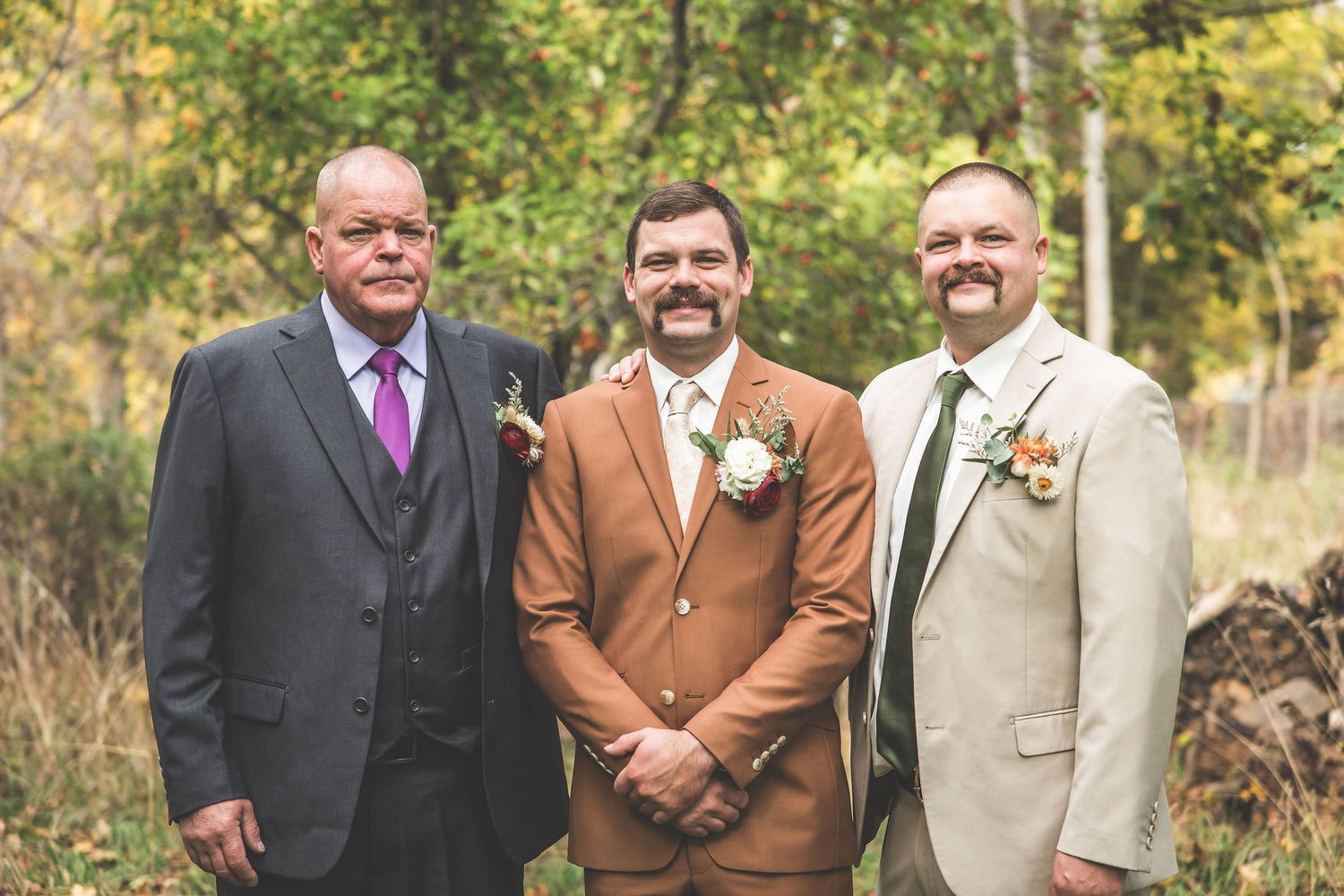 Group of men in formal suits pose together outdoors during fall wedding.
