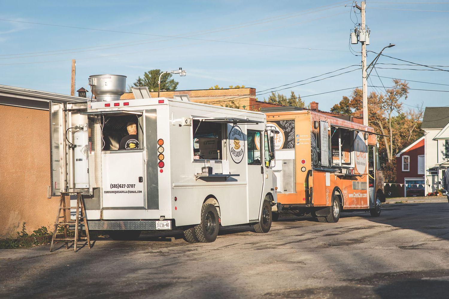 Row of white food trucks parked in a lot against blue sky and brick buildings.