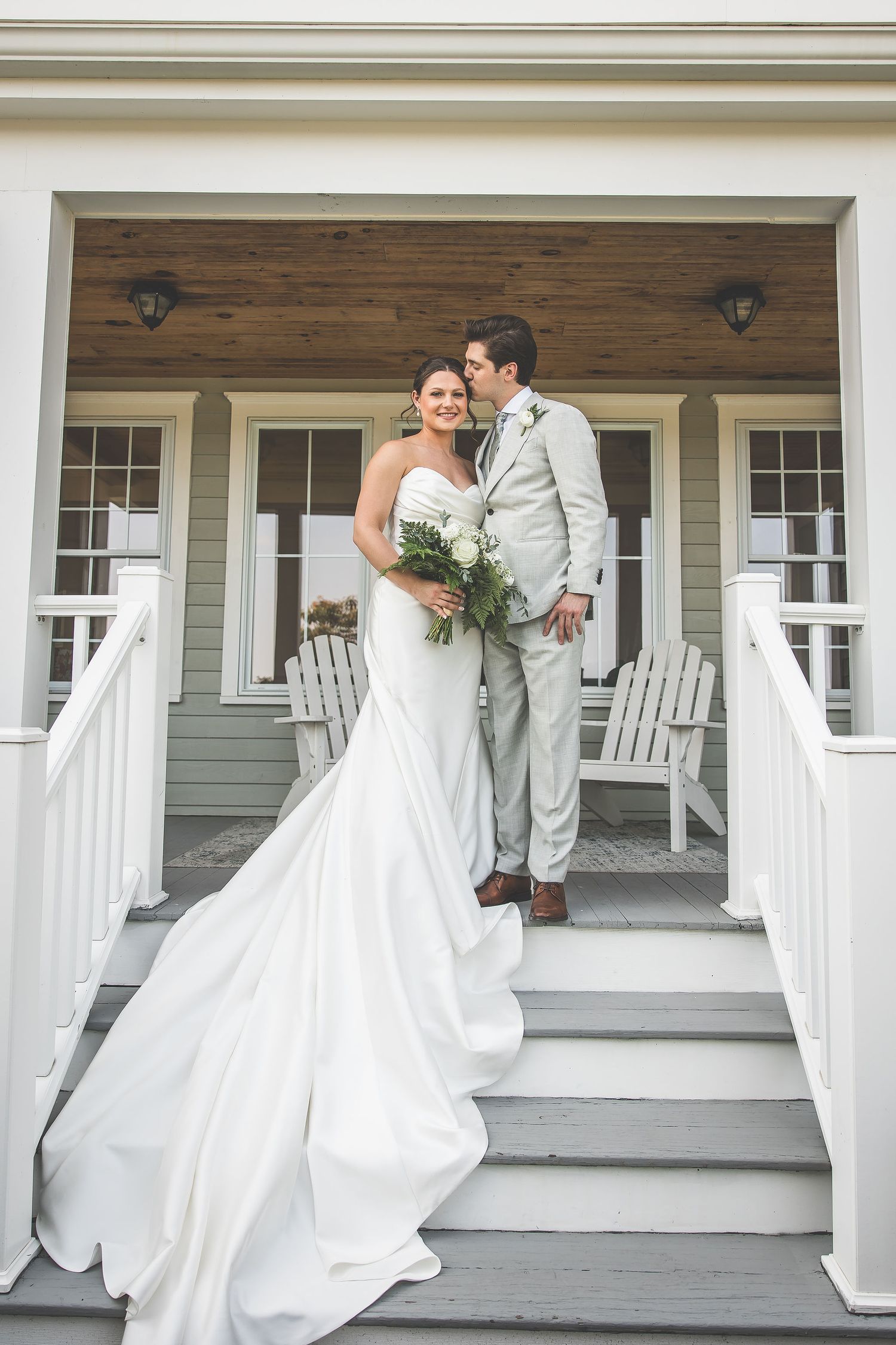 A couple shares an intimate moment on a white porch with classic columns and railing.
