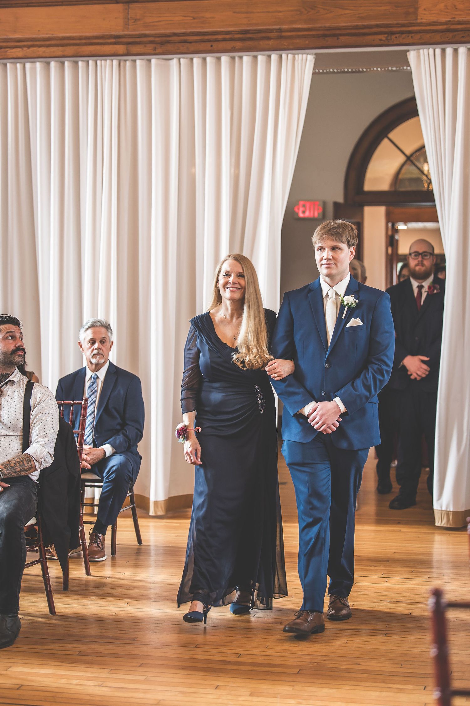 Wedding ceremony attendees standing in formal attire against white curtained backdrop.