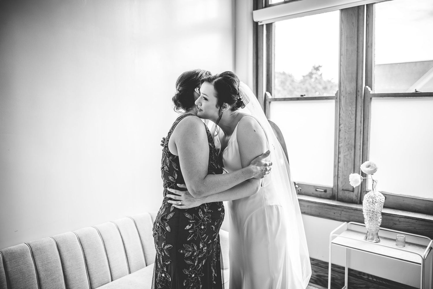 Black and white portrait of two people in wedding attire sharing a tender moment by a window.