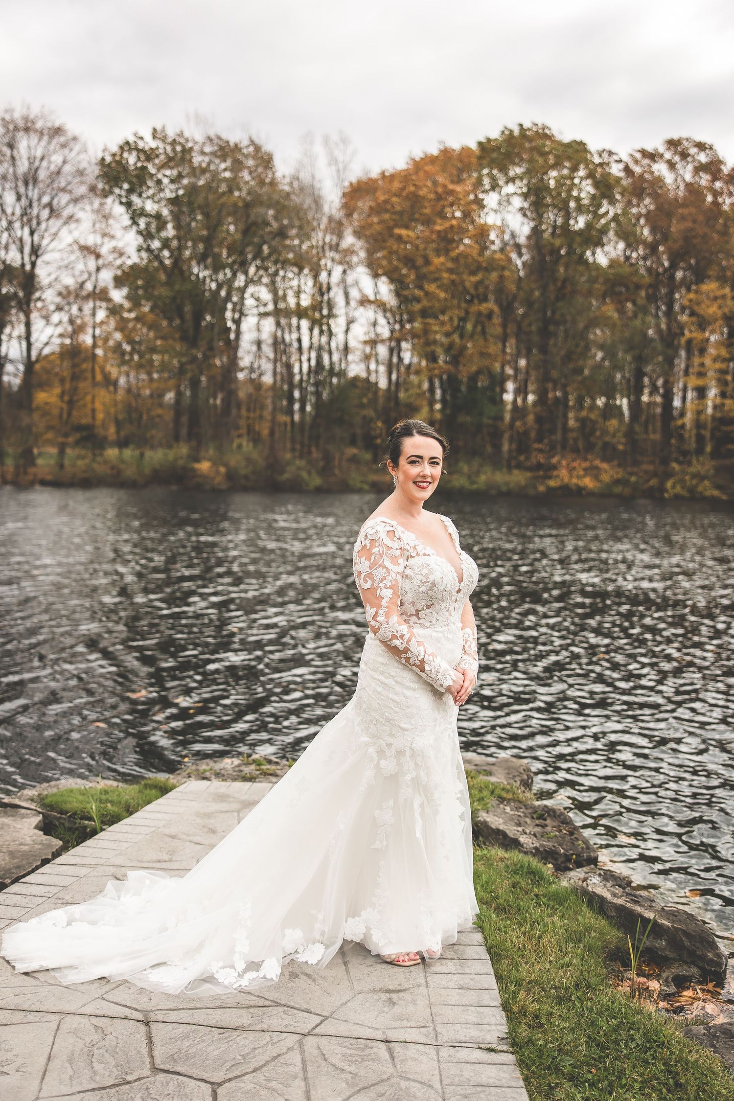 Bride in elegant lace wedding dress poses on stone path beside autumn lake.