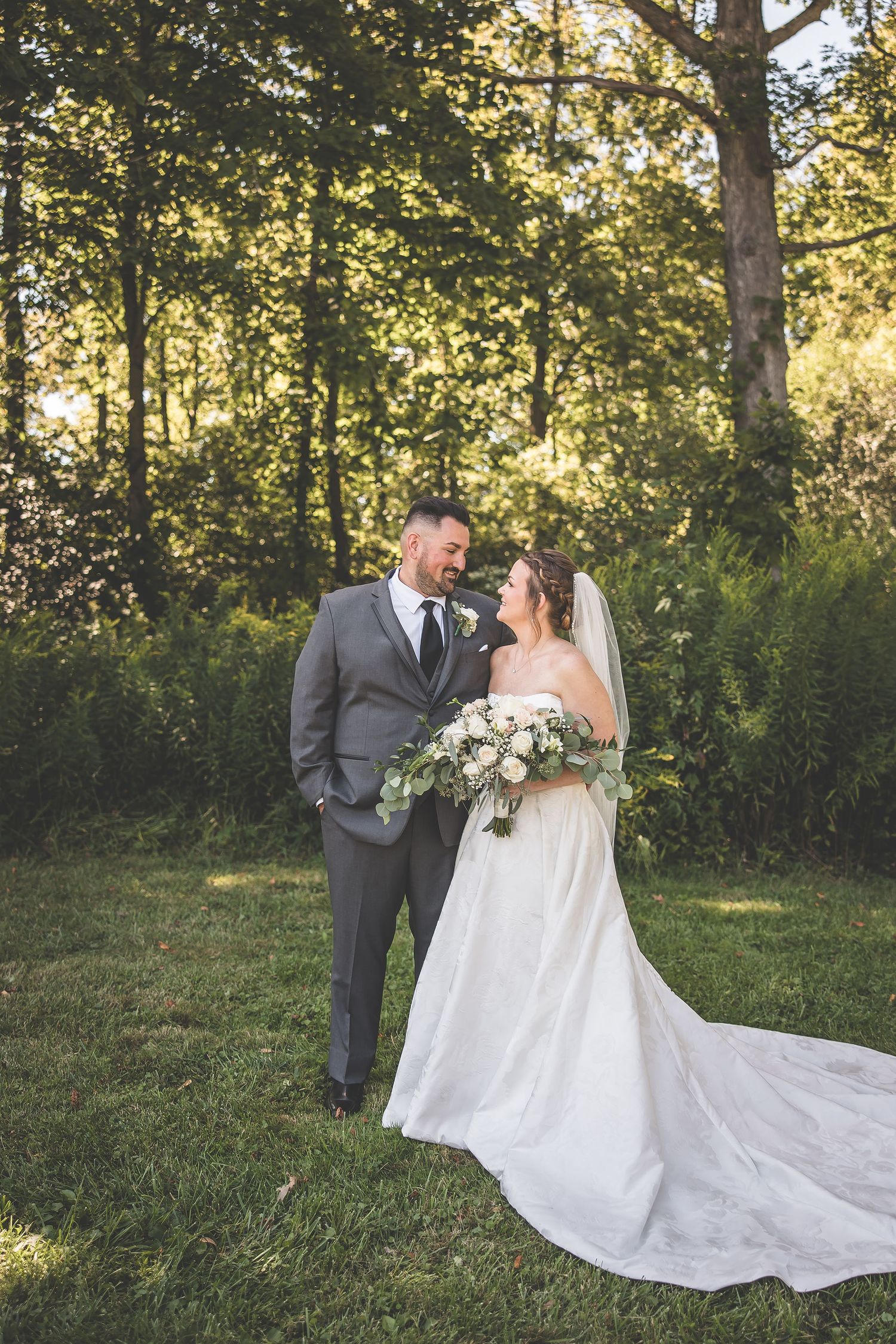 Romantic outdoor wedding portrait with bride's long train flowing on grass beneath tall trees.