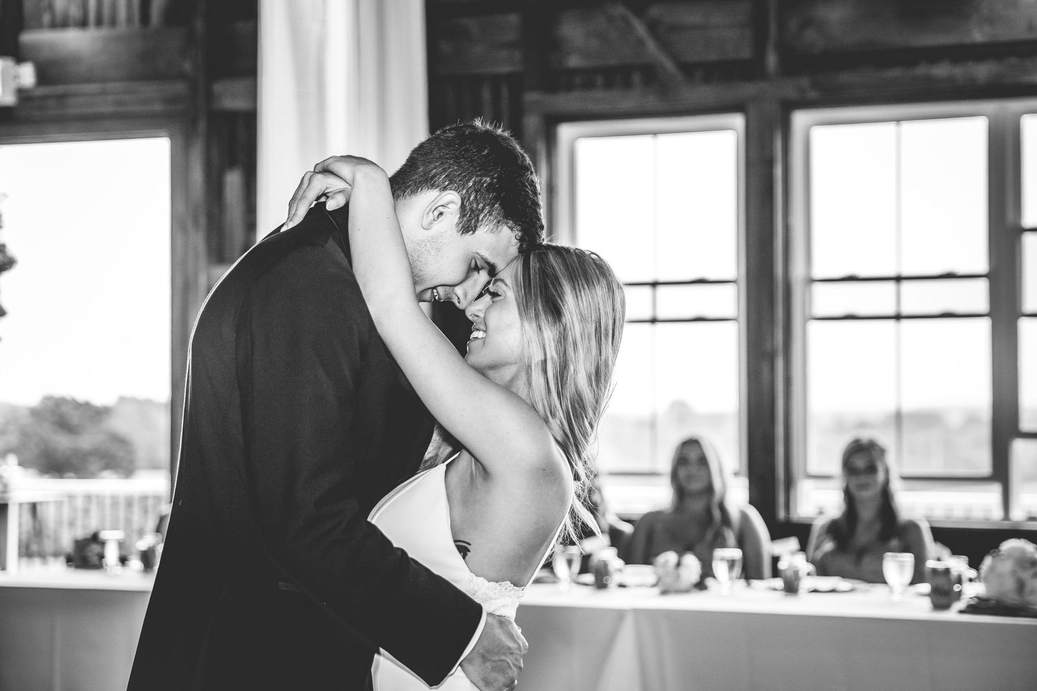 A romantic black and white wedding dance moment in a rustic venue with windows and guests in the background.