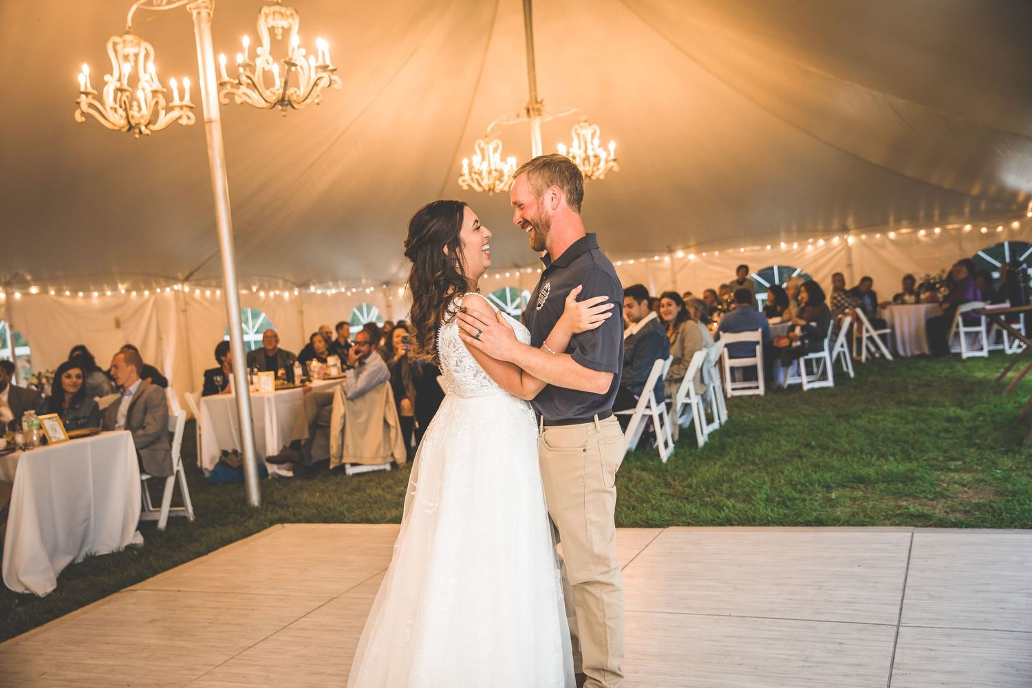 Wedding reception tent glows warmly with string lights and chandeliers as guests celebrate.