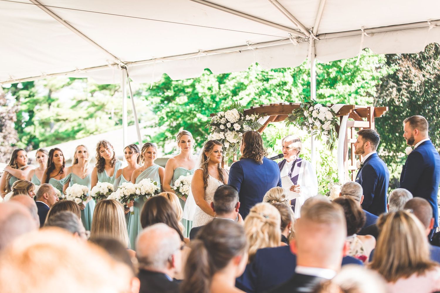 Wedding guests seated under tent canopy with lush green trees in background.