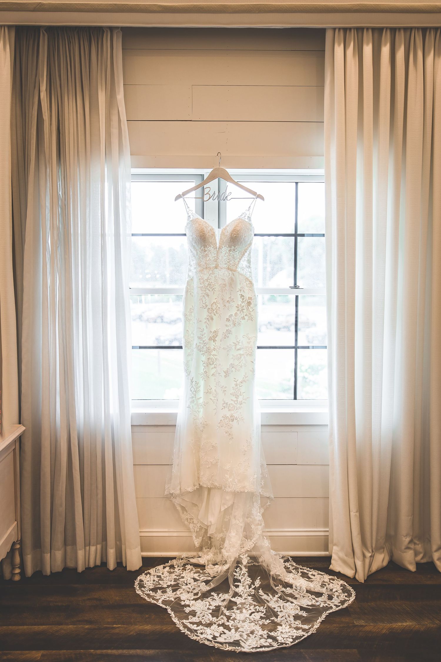 Elegant white lace wedding dress with intricate detailing hangs by a window in a bridal suite.