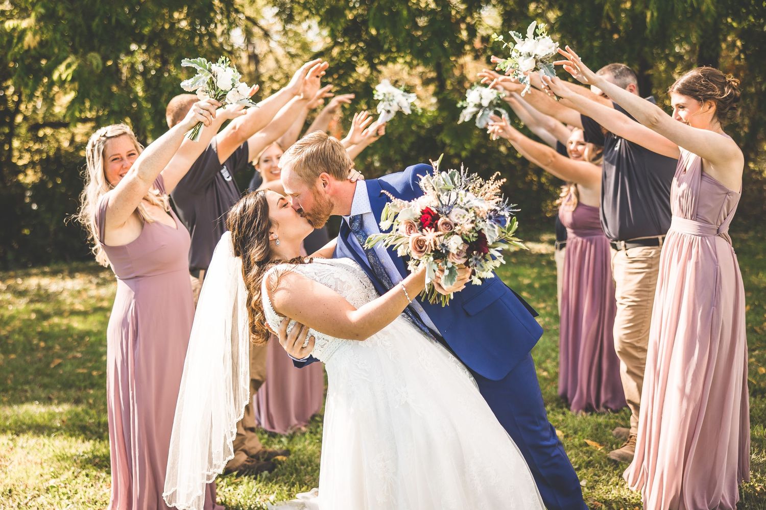 Wedding party creates arch with raised arms as couple shares romantic kiss under trees.