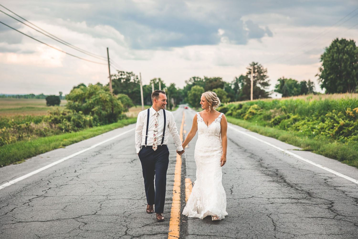 Newlyweds holding hands while strolling down a rural road surrounded by green fields.