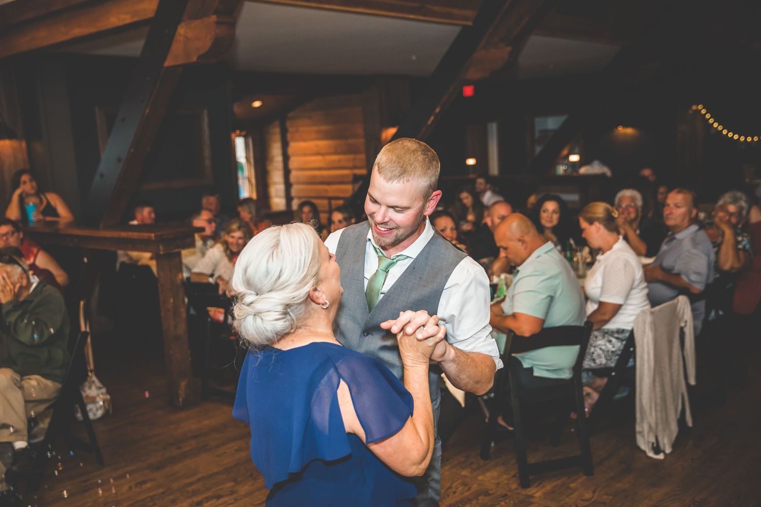 Wedding guests dancing and celebrating on a wooden dance floor at an indoor reception venue.