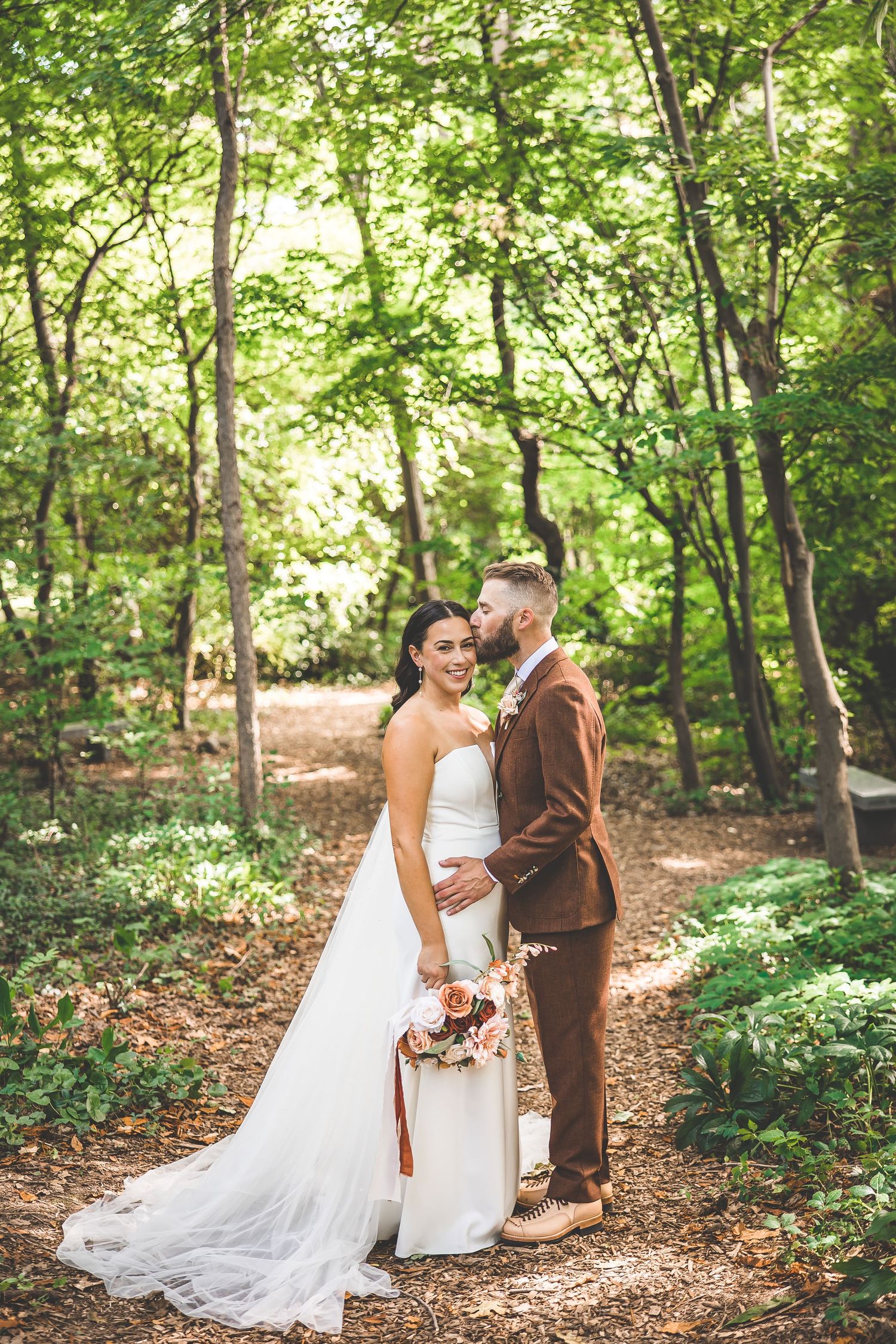Wedding couple embraces on a wooded trail with dappled sunlight filtering through the trees.