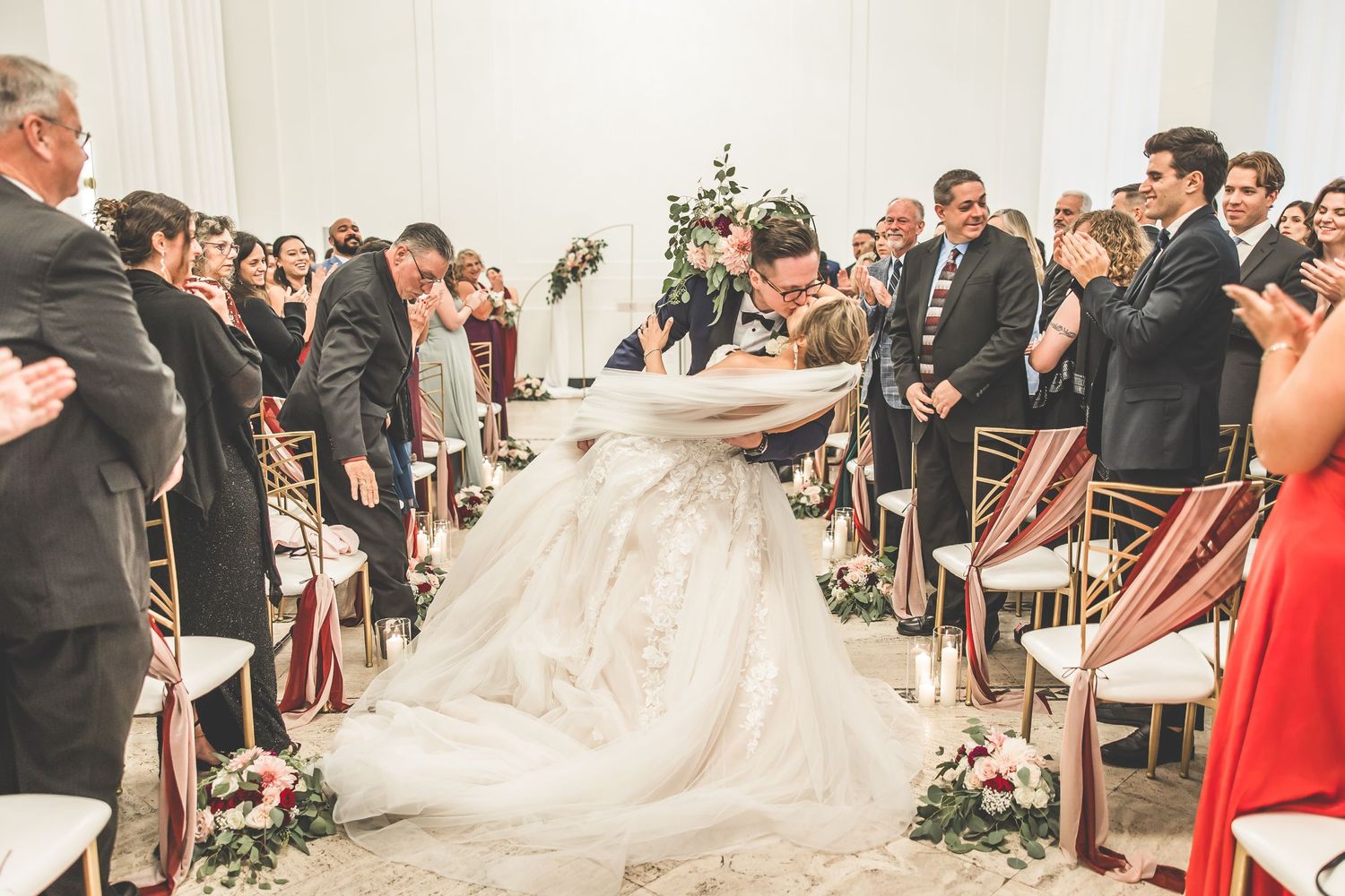 Long flowing wedding dress train trails behind couple as they exit ceremony with guests looking on.