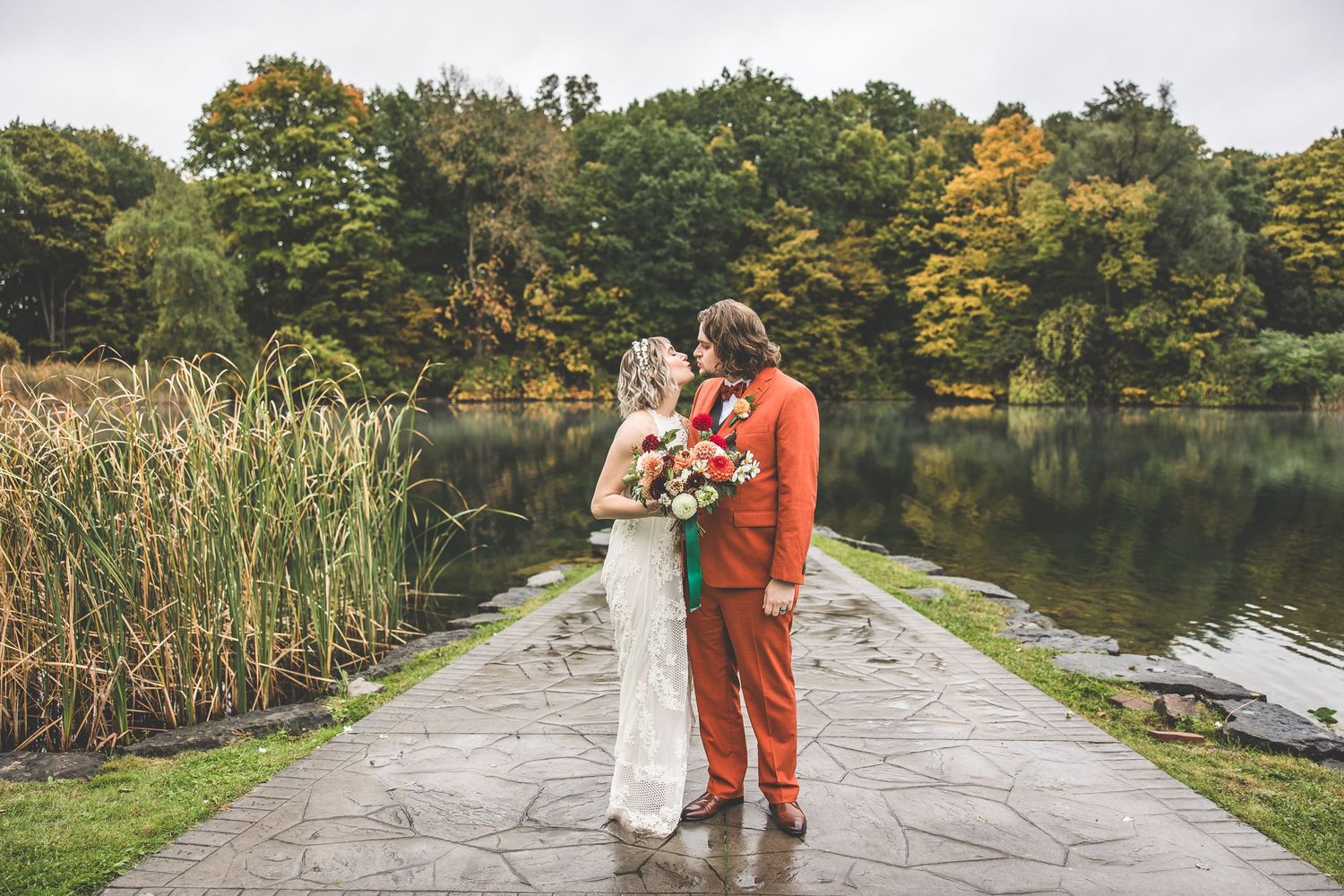 Newlyweds share intimate moment on lakeside path surrounded by fall colors and tall reeds.