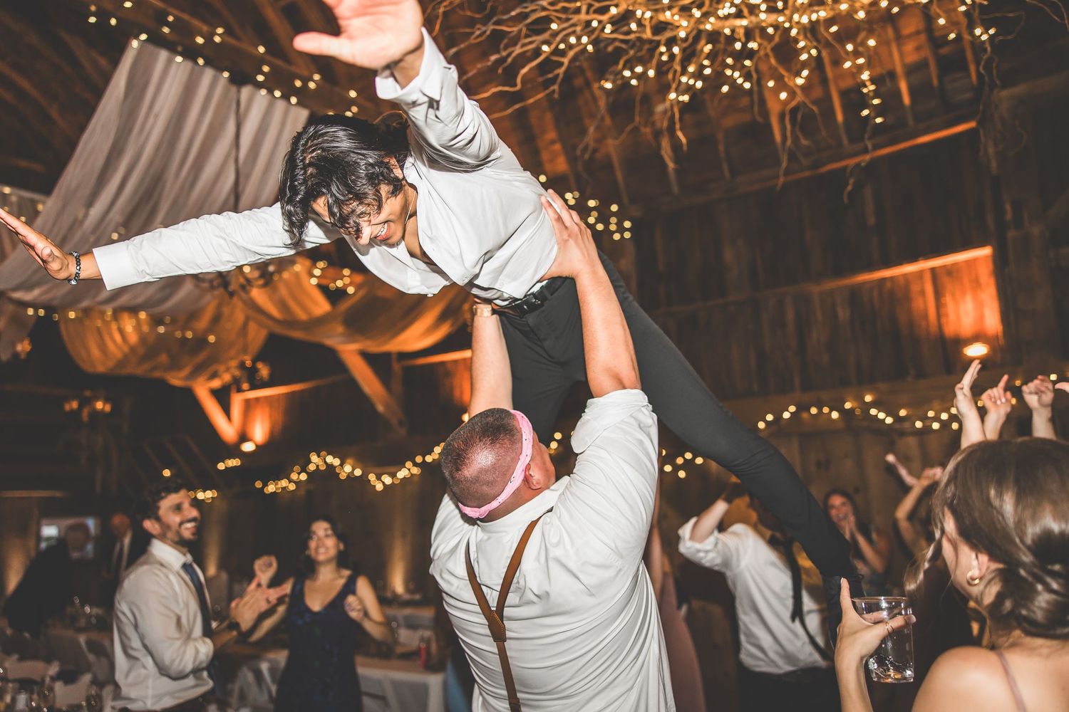 Wedding celebration with guests dancing under string lights in a rustic indoor venue.