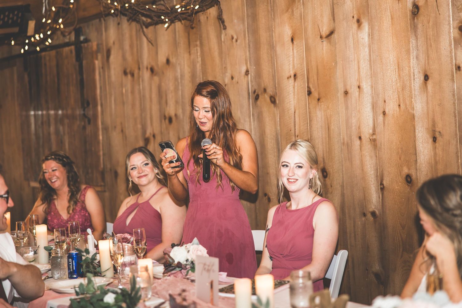 Bridesmaids in burgundy dresses give speeches during reception in rustic barn venue.