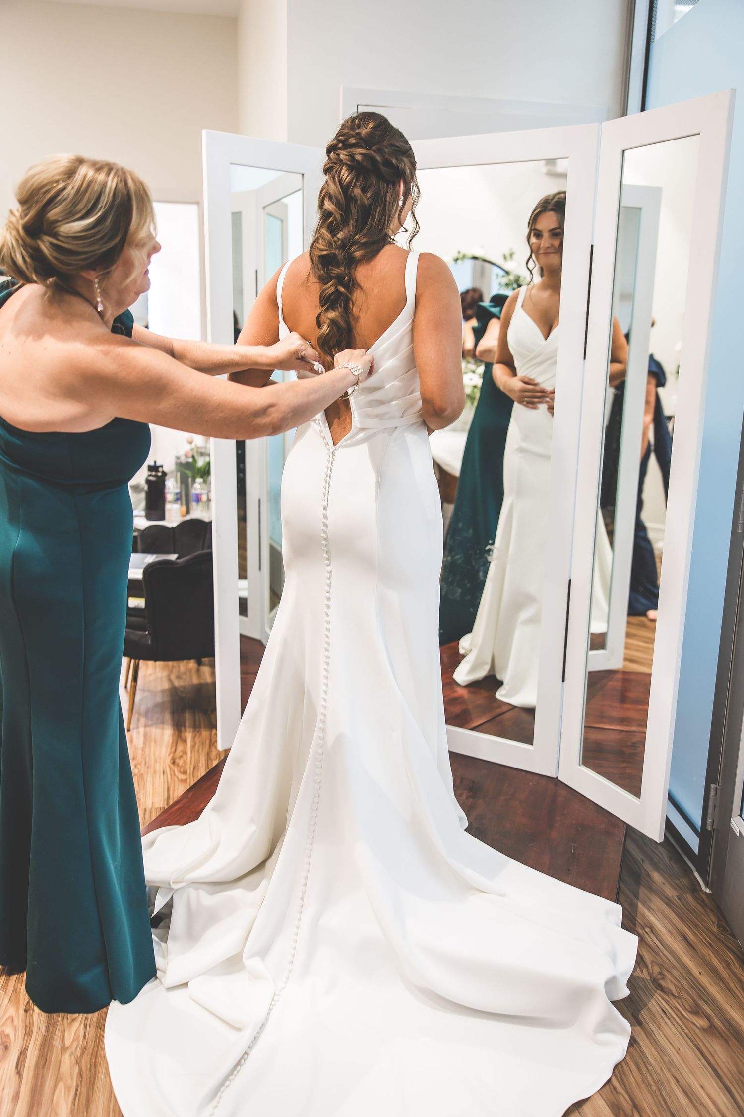Women in teal bridesmaid dresses help adjust a white wedding gown in a room with mirrors.
