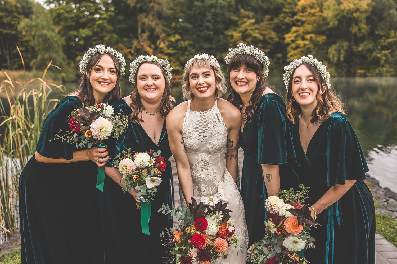 Wedding party in emerald velvet dresses and white gown poses with floral bouquets by lake.